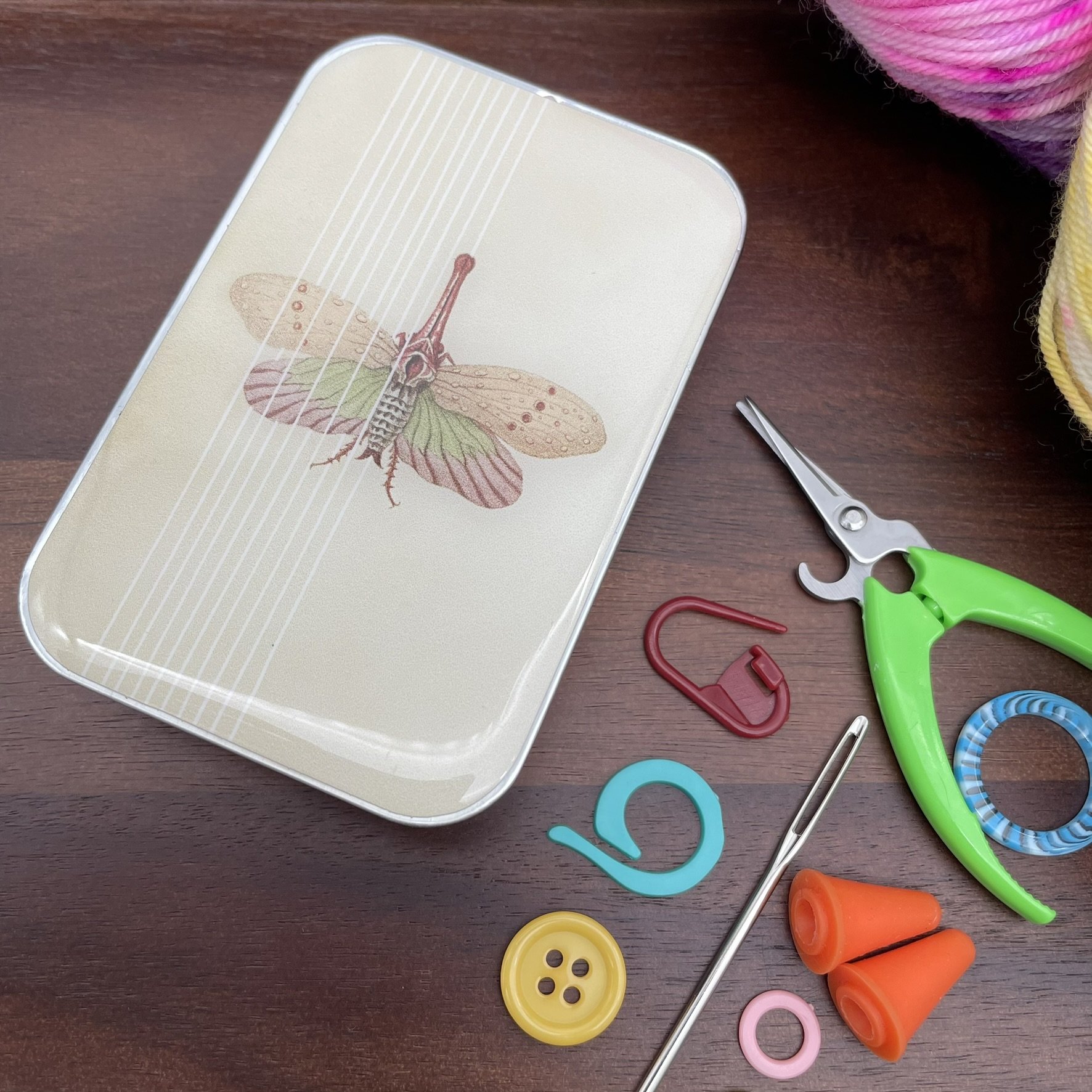 Spring Moth design notions tin by Firefly Notes shown with knitting accessories on wooden surface.