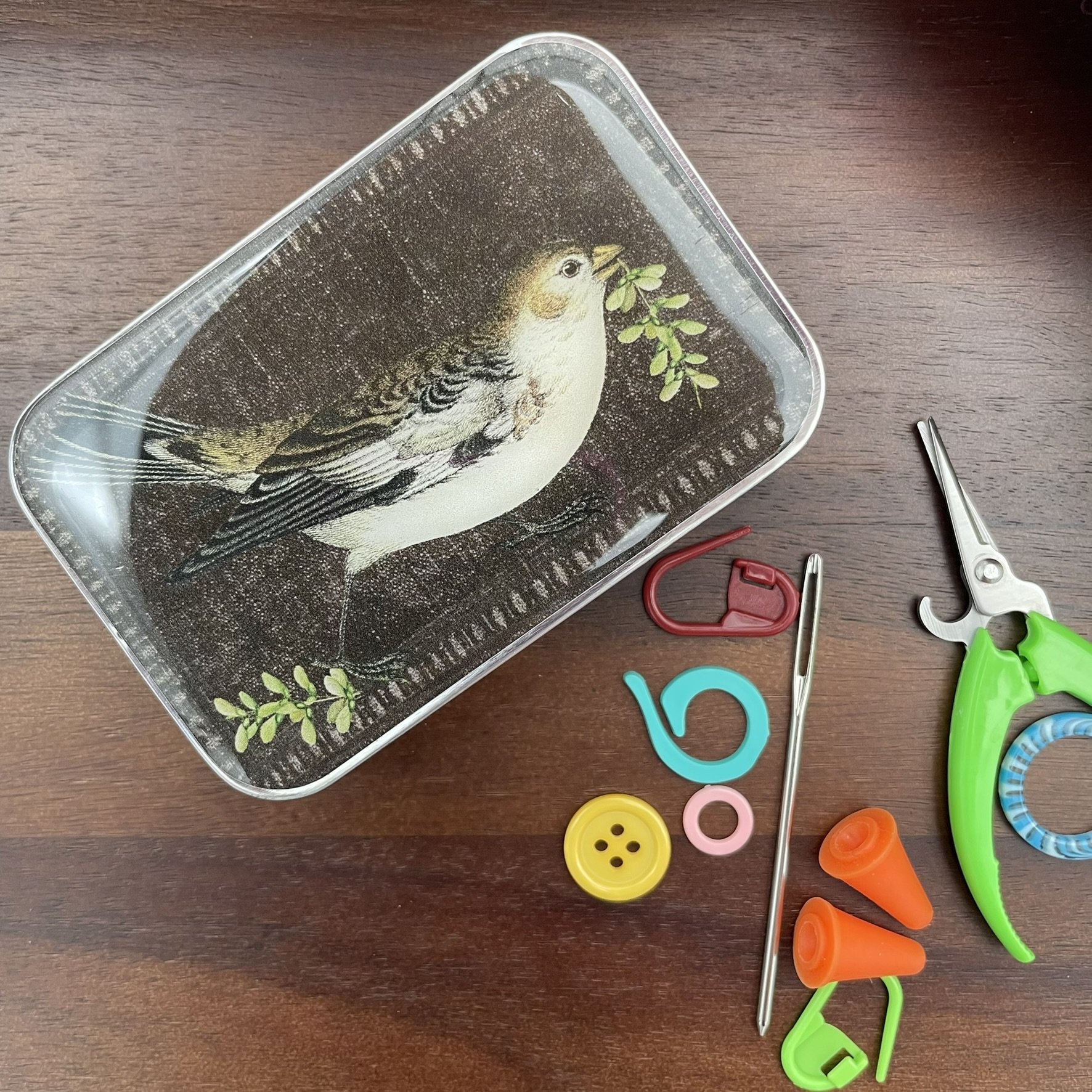 Top view of a bird-themed notions tin, showcasing a bird illustration on a dark background, with knitting notions nearby.