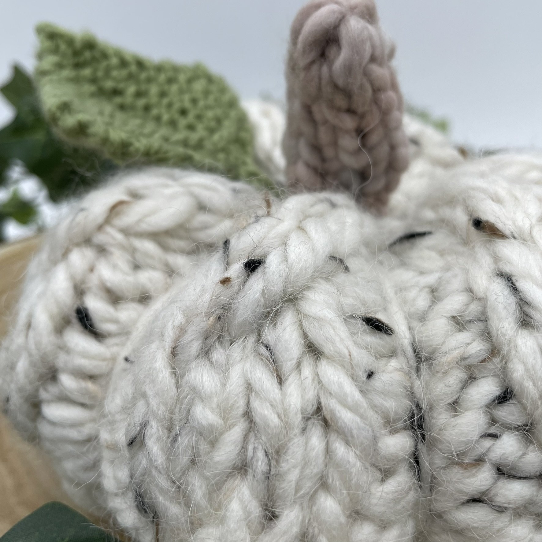 Hand holding a hand-knit white cable pumpkin to show size reference, featuring a green leaf and beige stem.