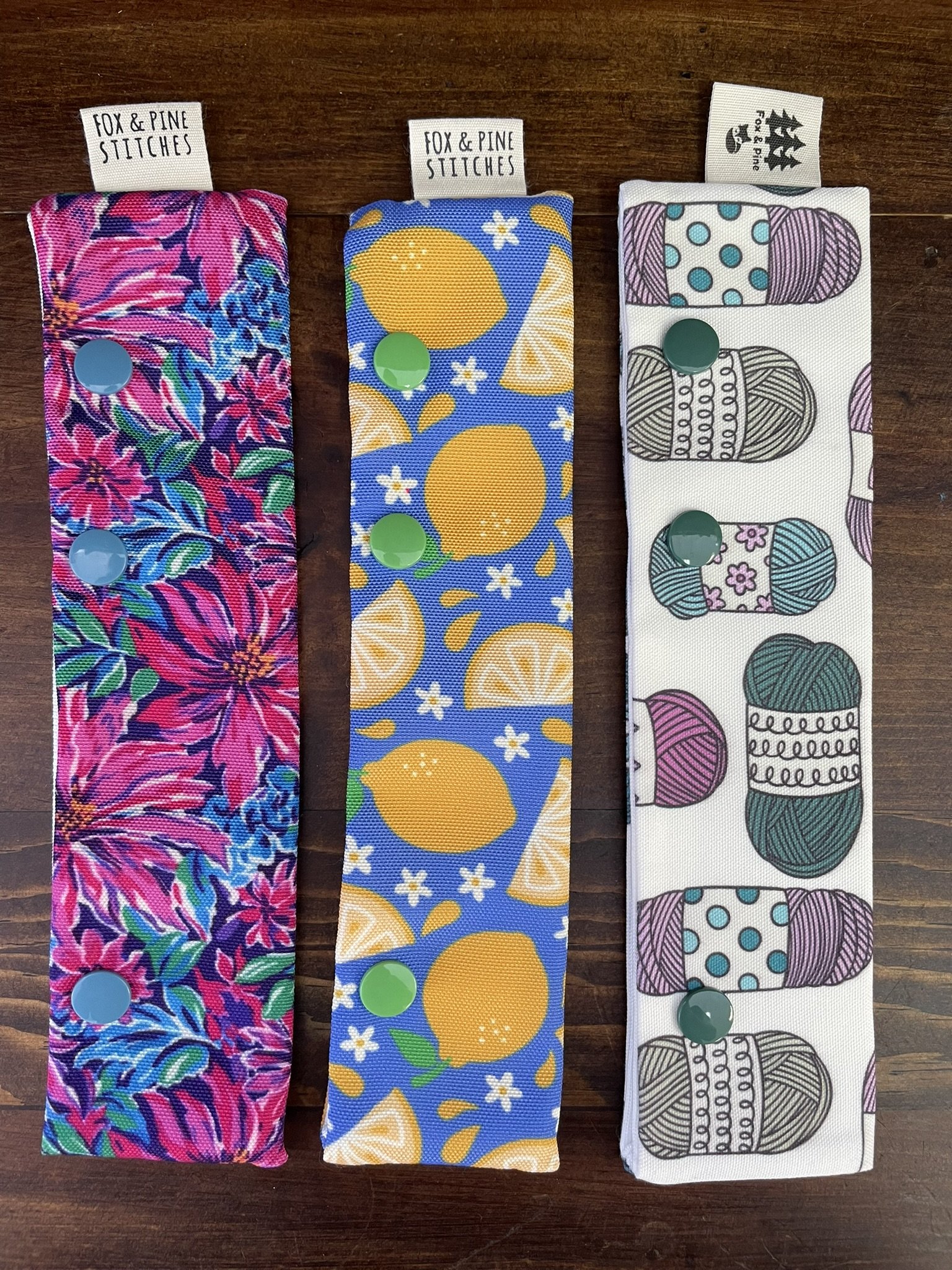 Fox & Pine double-pointed needle holders with colorful designs featuring lemons, floral patterns, and yarn prints, displayed on a wooden table.
