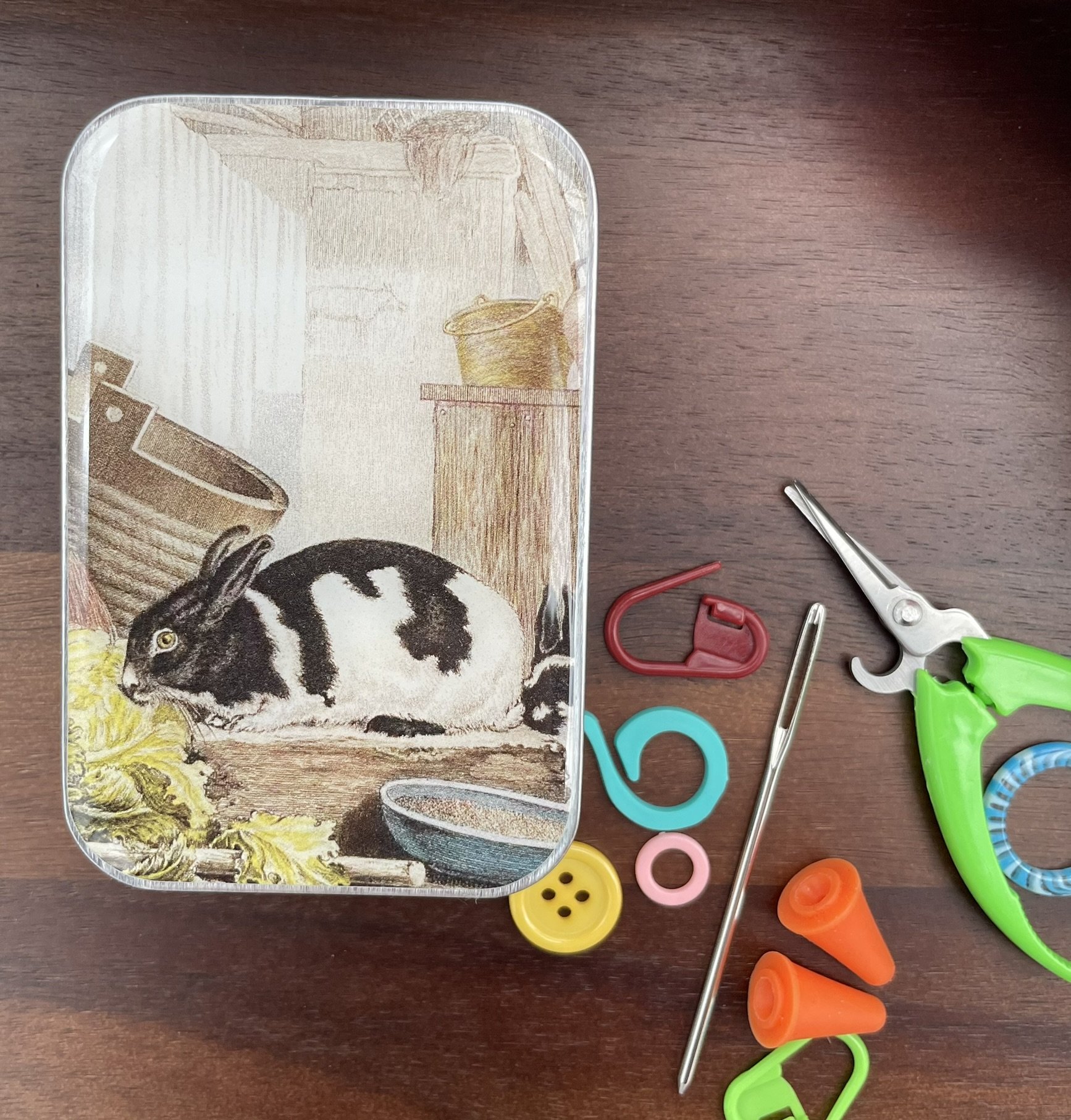 Top view of notions tin with a vintage rabbit illustration next to knitting tools.