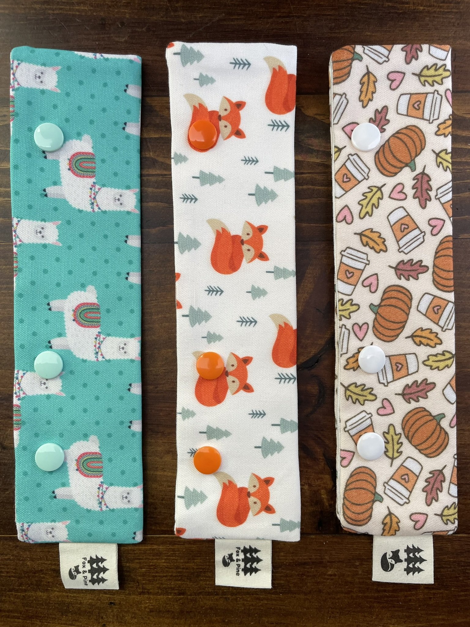 Fox & Pine double-pointed needle holders with designs featuring llamas, foxes, and fall motifs, placed on a wooden background.