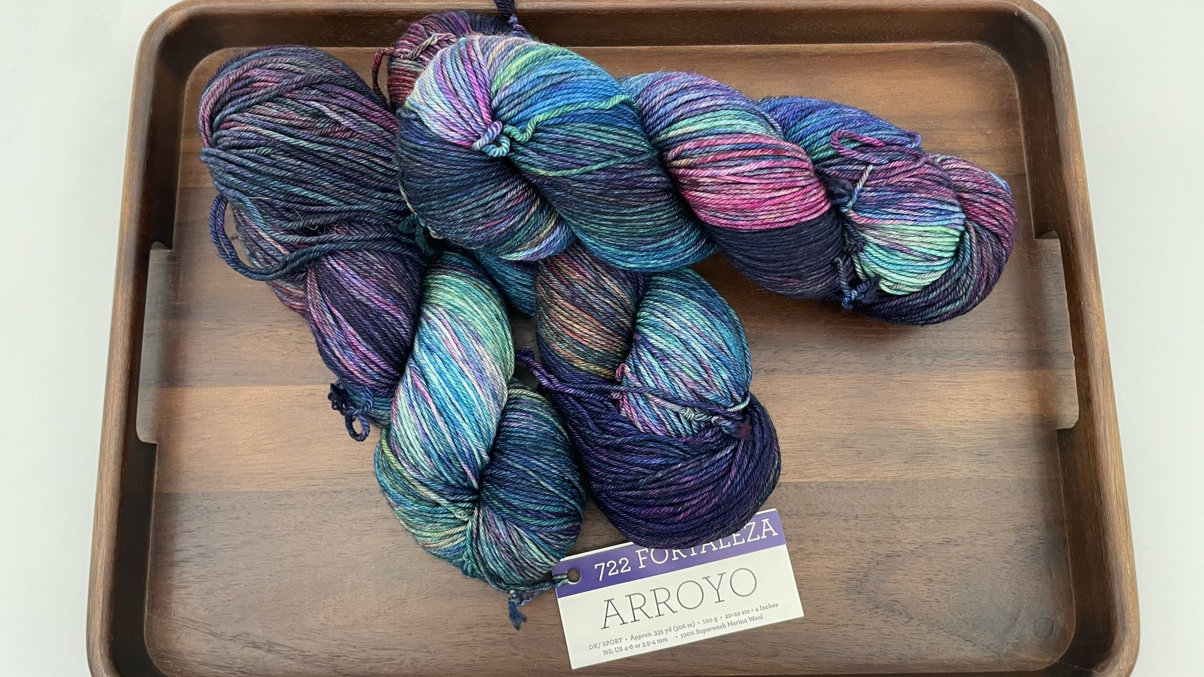 Pile of Malabrigo Arroyo Fortaleza yarn in DK/Sport weight, showcasing vibrant colors on a wooden tray.
