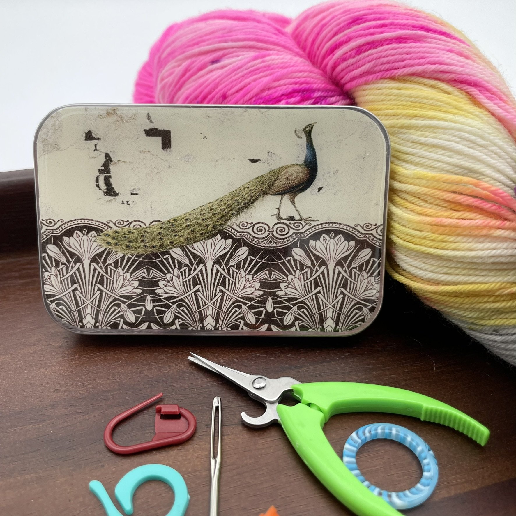 Upright notions tin with a vintage peacock illustration alongside knitting accessories.