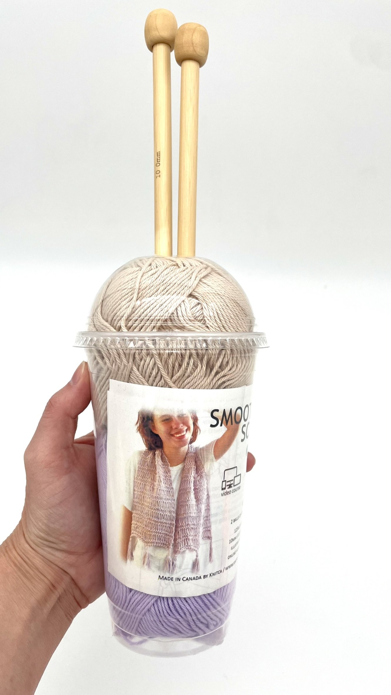 DIY Smoothie Scarf Knitting Kit by Knitca showing tan and purple yarn held in hand for size reference, with wooden knitting needles.