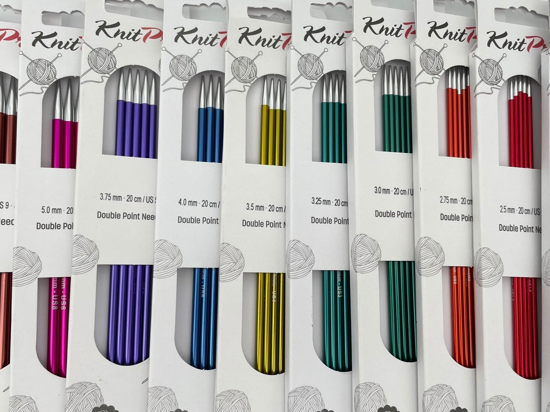 Colorful double-point knitting needles in various colors and sizes packaged in white boxes with a ball of yarn graphic.