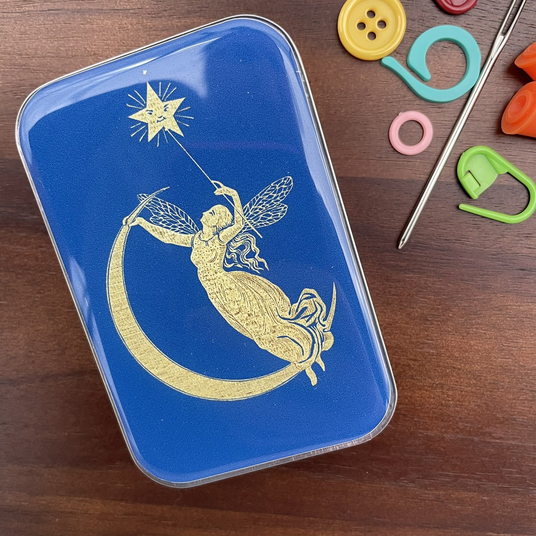 Top view of a blue fairy design notions tin featuring a gold fairy reaching for a star, with yarn and knitting tools around.