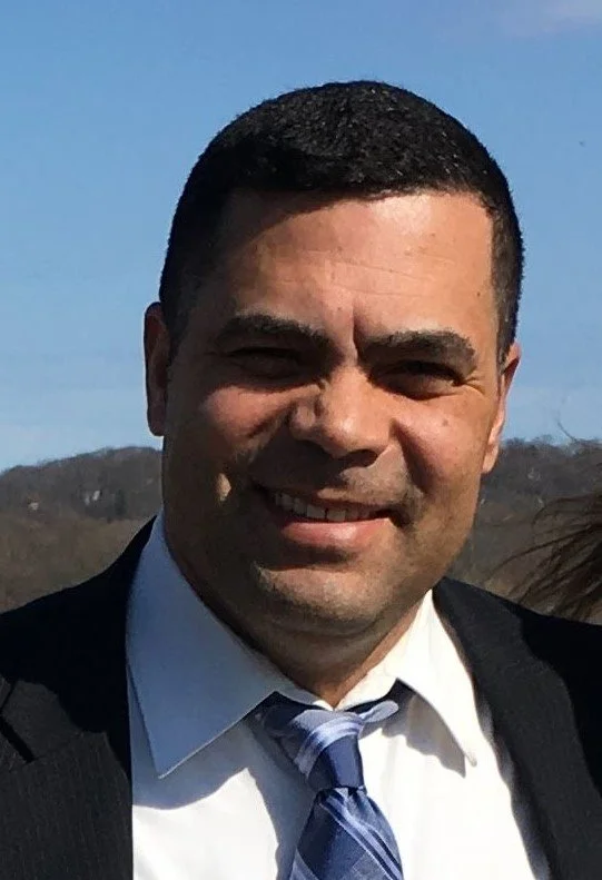 A man smiling outdoors, wearing a suit, white shirt, and a blue tie, with a hilly landscape in the background.