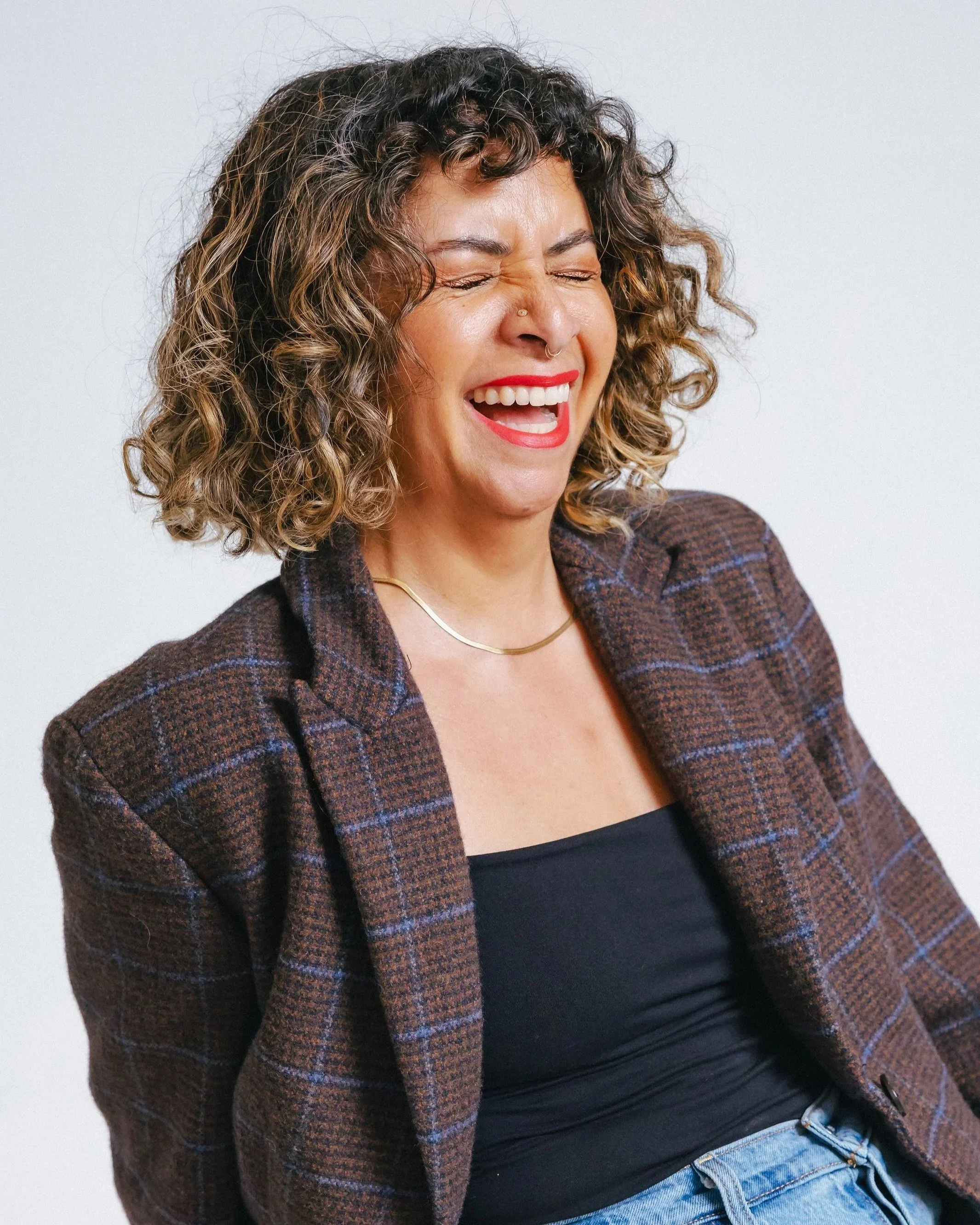A woman with curly hair and a septum piercing laughing, wearing a plaid blazer over a black top and blue jeans, against a plain white background.