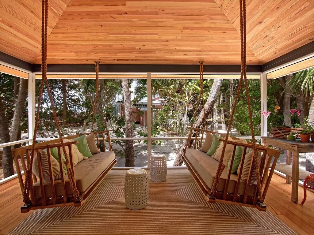 Two wooden porch swings with cushions and pillows are hung from a wooden ceiling with thick ropes, facing large windows overlooking a garden with trees and greenery.