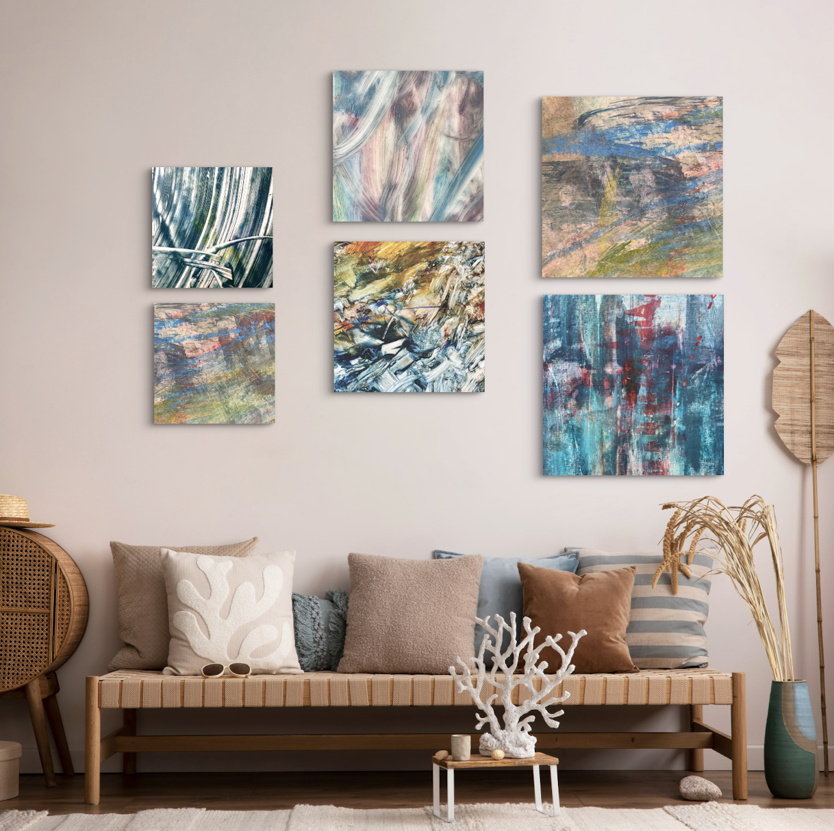 A living room with a beige sofa decorated with a variety of cushions and a small table with coral decor in front. The wall above features a collection of six abstract paintings in different sizes and colors. There is a tall green vase with dried plants on the right side and a rattan fan or sculpture on the left side.