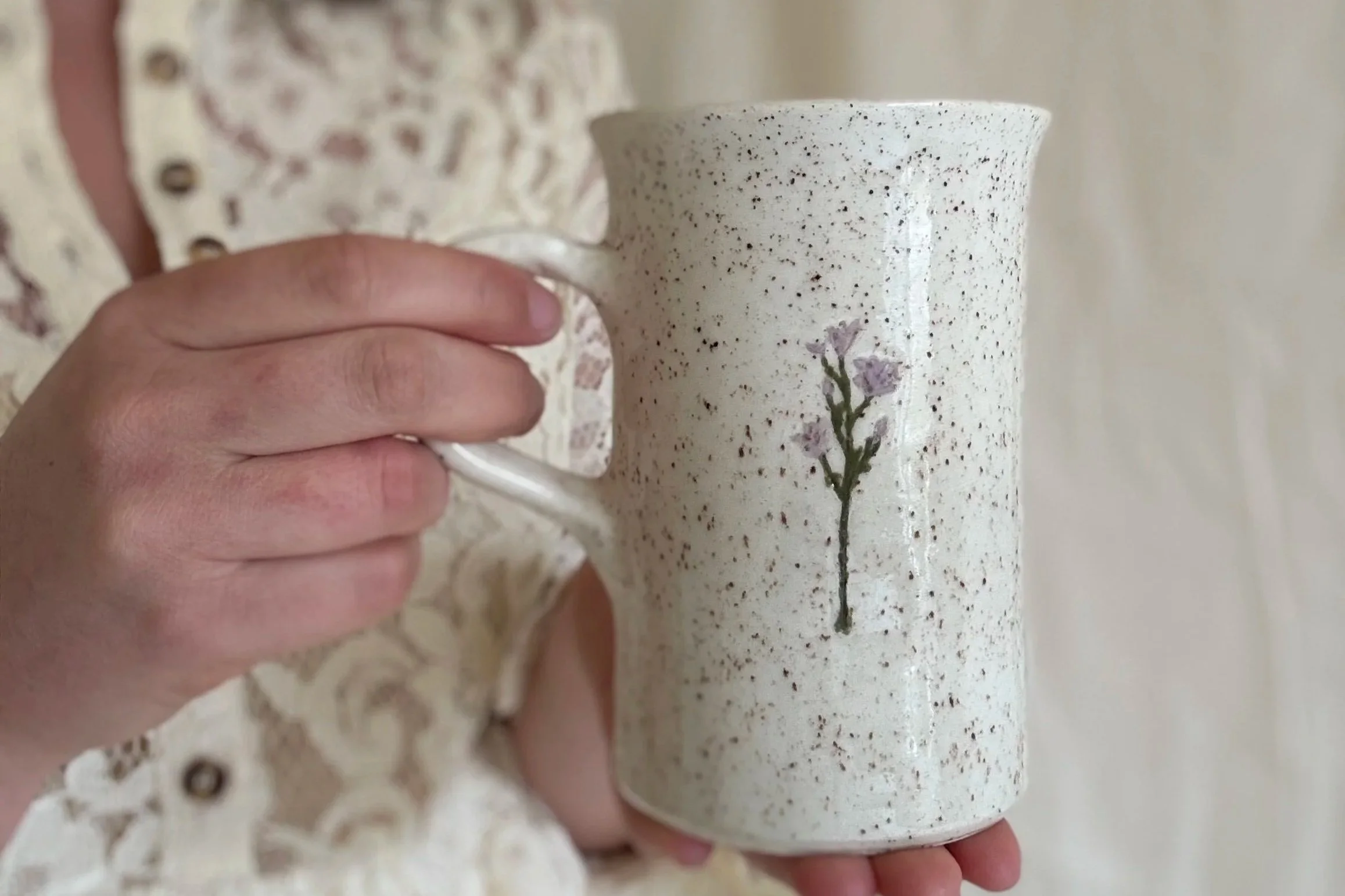 A hand holding a speckled ceramic mug with a small painted purple flower and green leaves design.