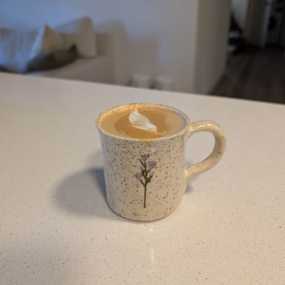 A speckled ceramic mug with a small purple flower design, filled with coffee topped with foam art, on a white countertop in a cozy home setting.
