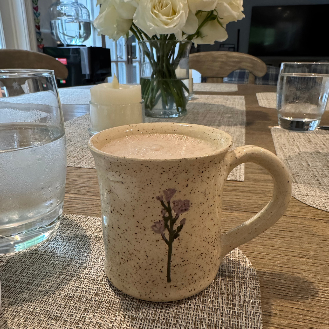 A ceramic mug with a floral design on a wooden dining table, filled with a frothy beverage. In the background, a glass of water, a vase of white roses, a lit candle, and other glasses are visible, with a window and a television in the background.