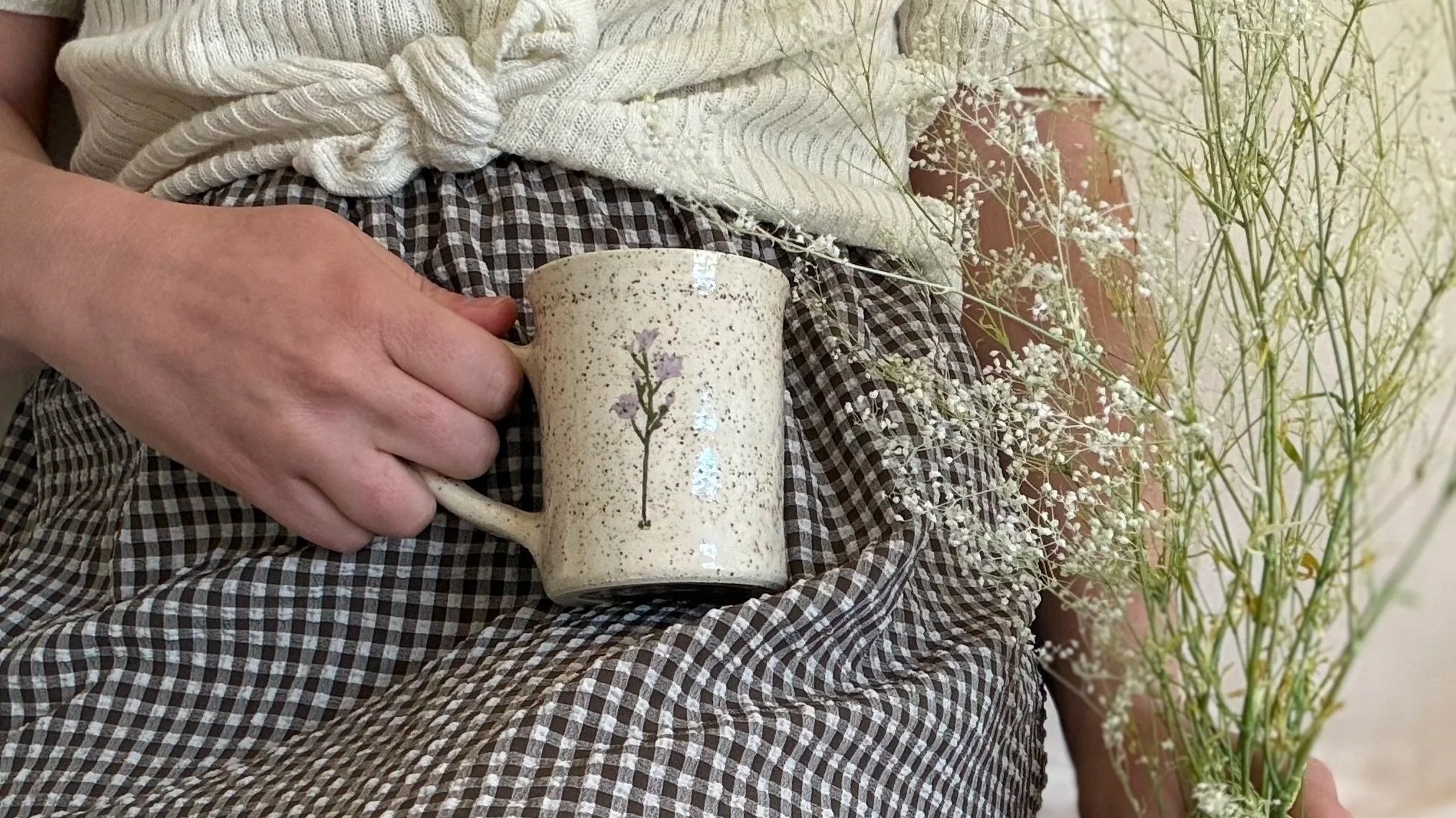 Person holding a speckled beige mug with a small flower illustration on it, sitting near plants with tall, white, delicate flowers.