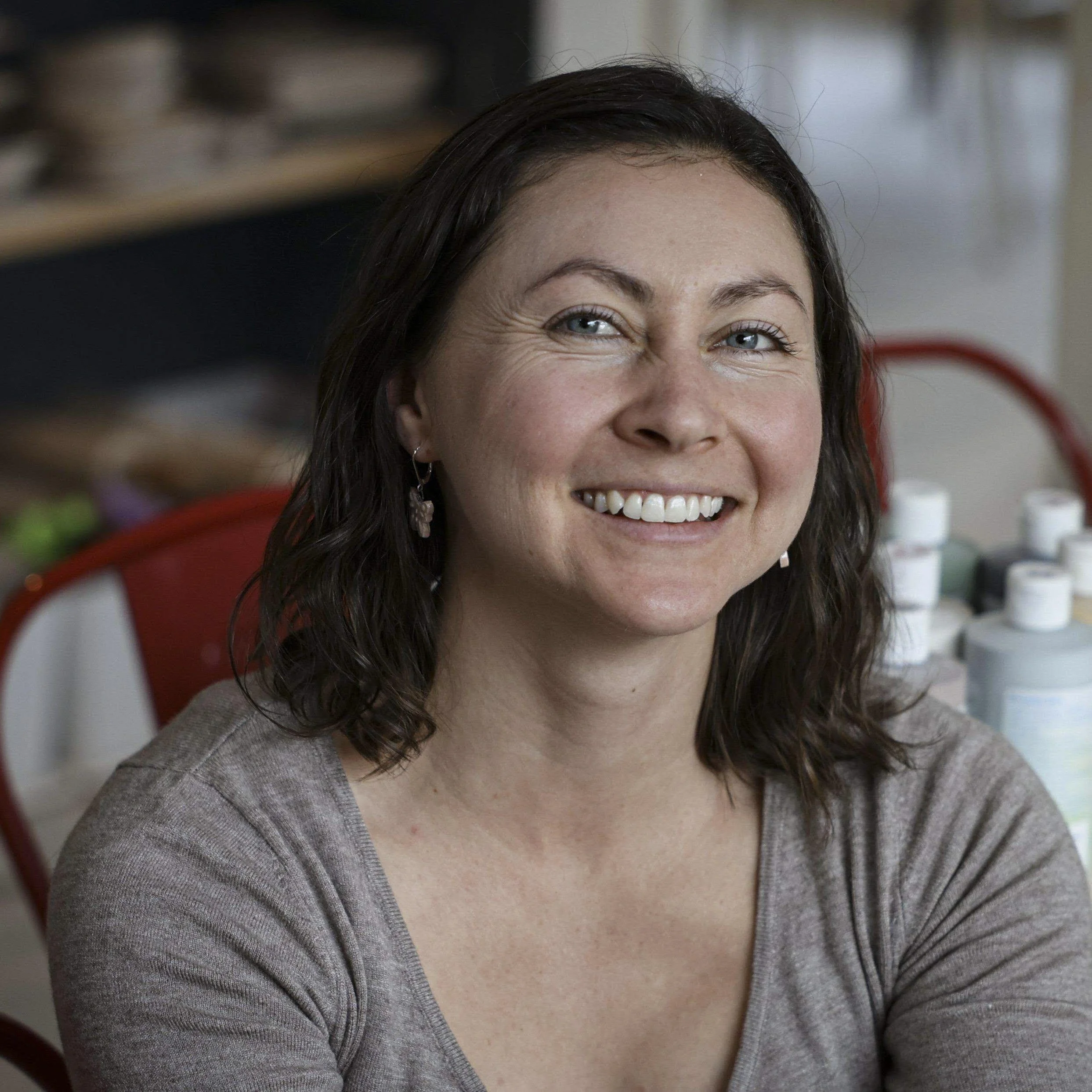 Close-up of a smiling woman with blue eyes and dark brown hair, wearing a gray cardigan, in a room with shelves and objects in the background.