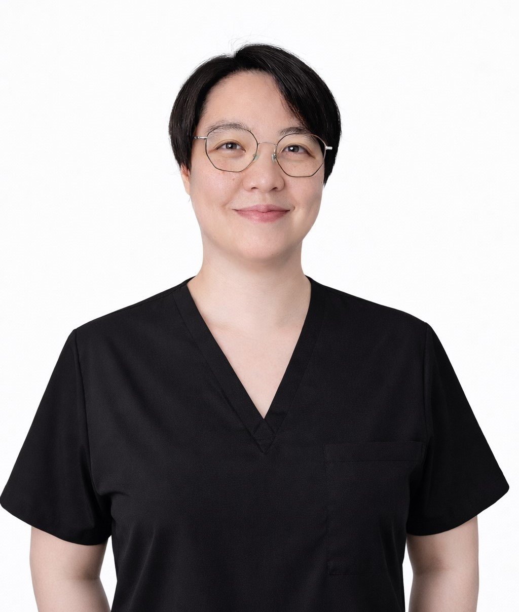 A woman with dark hair tied back, wearing a black top and a gold necklace, smiling against a white background.
