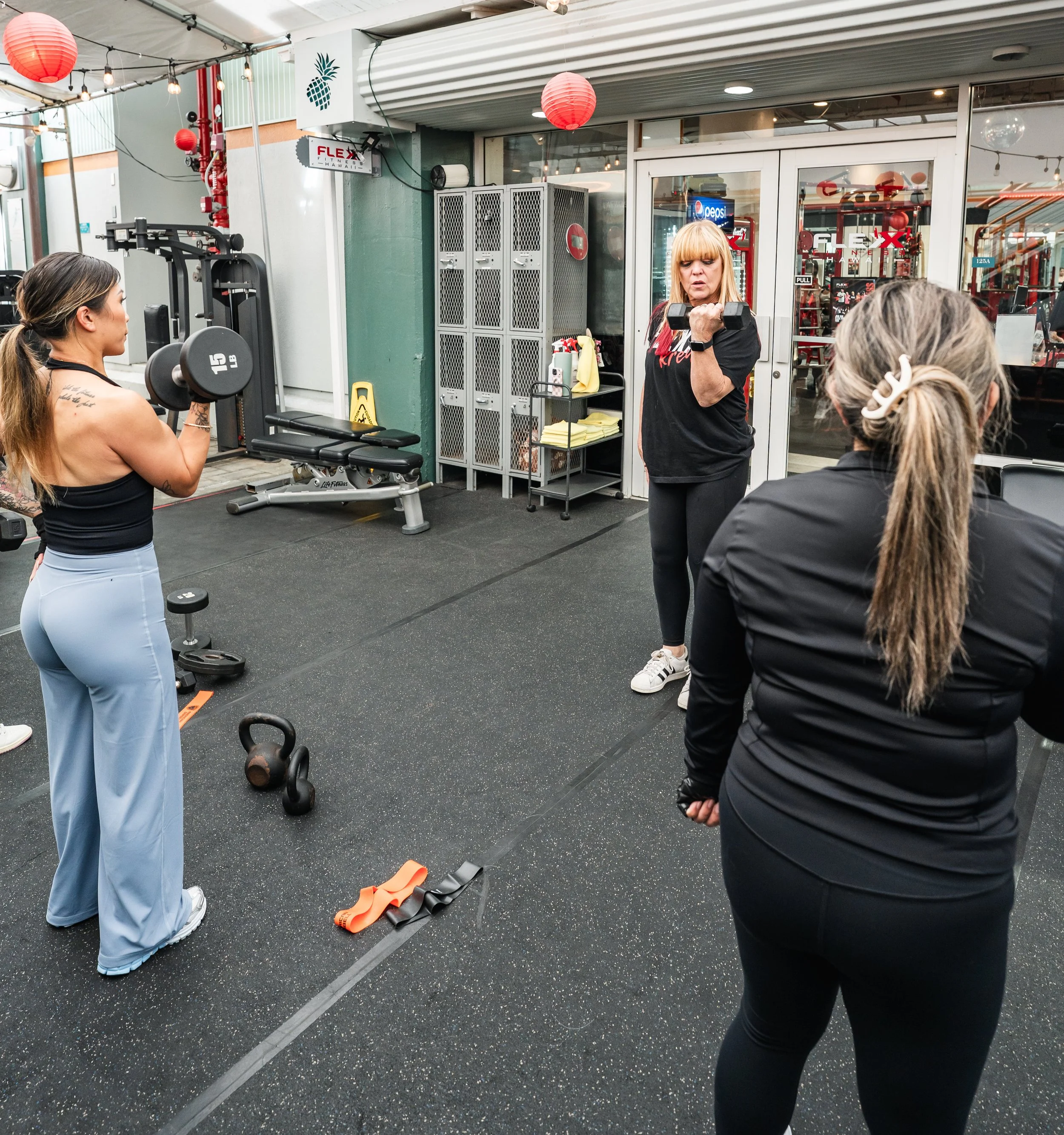 Women in workout gear training at a gym, with one woman holding a dumbbell, another listening, and a third with gray hair. Gym equipment, lockers, and red lanterns decorate the space.
