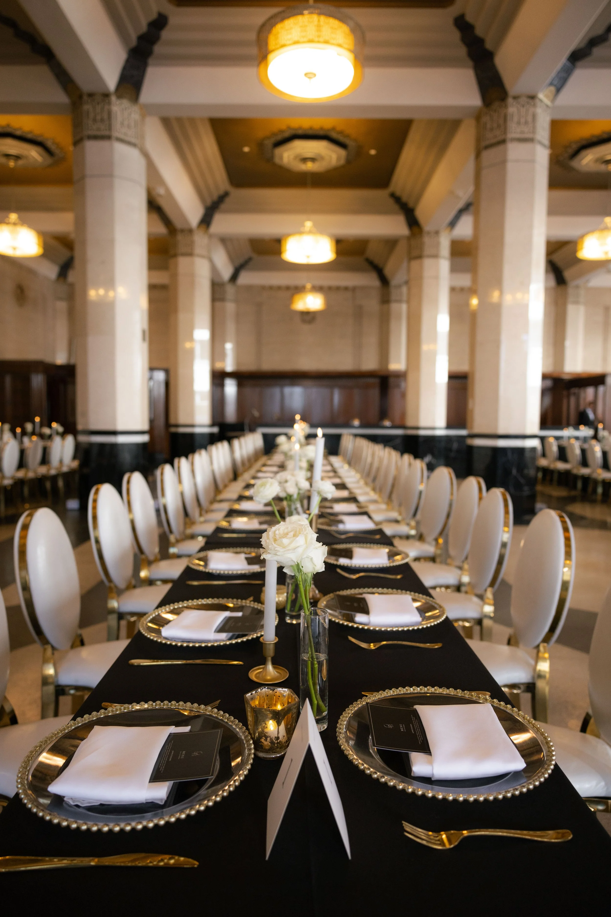 Elegant banquet table set for a formal event with black tablecloth, white napkins, gold cutlery, white roses, candles, and place settings, in a grand hall with black and white columns and warm lighting.