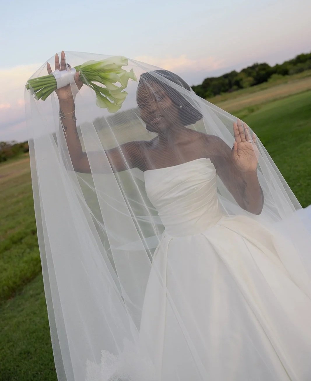 A woman in a wedding dress and veil holds a bouquet of white flowers behind a veil outdoors on a grassy field with trees and a colorful sky in the background.