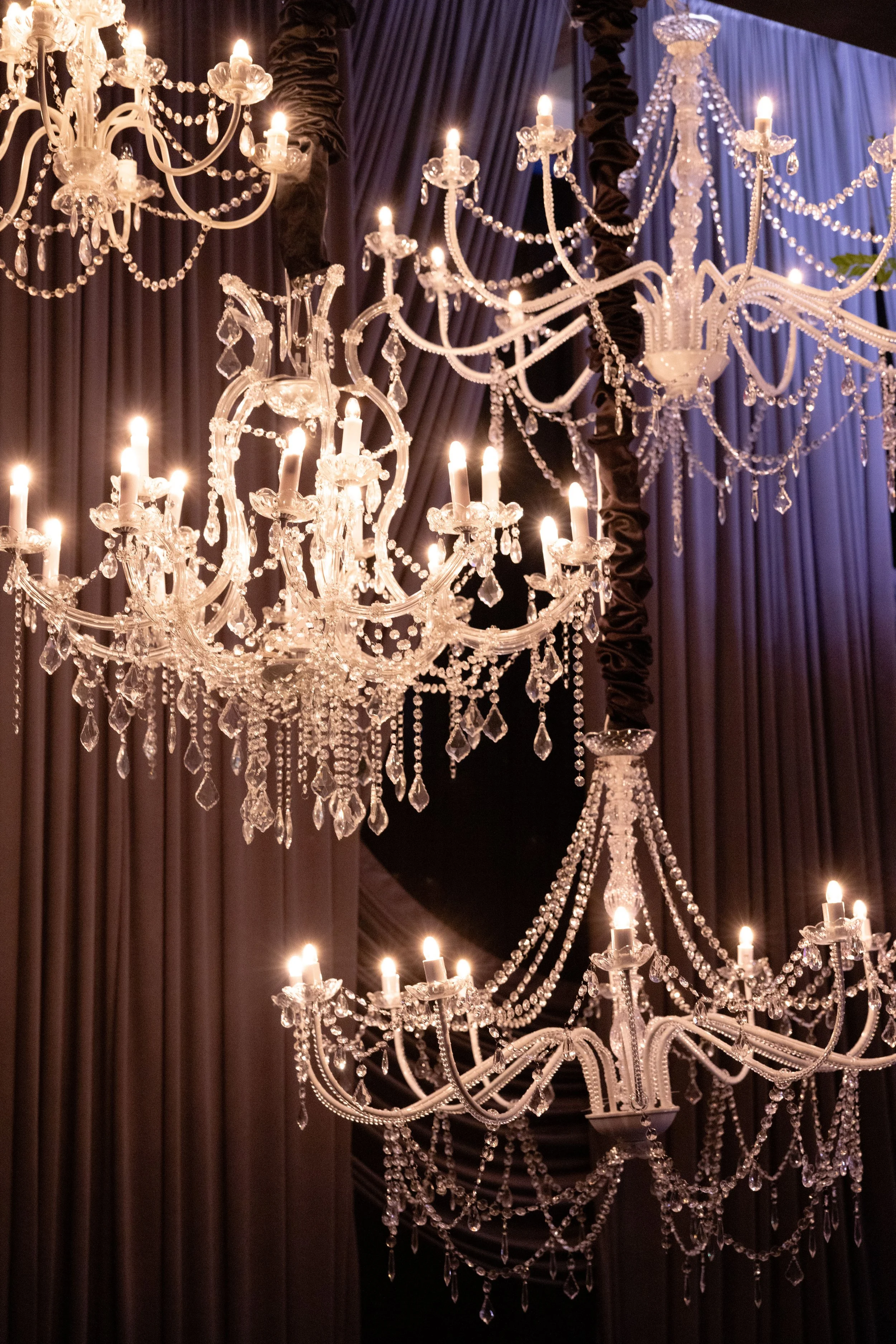 Elegant crystal chandeliers hanging from ceiling with dark drapery in the background.