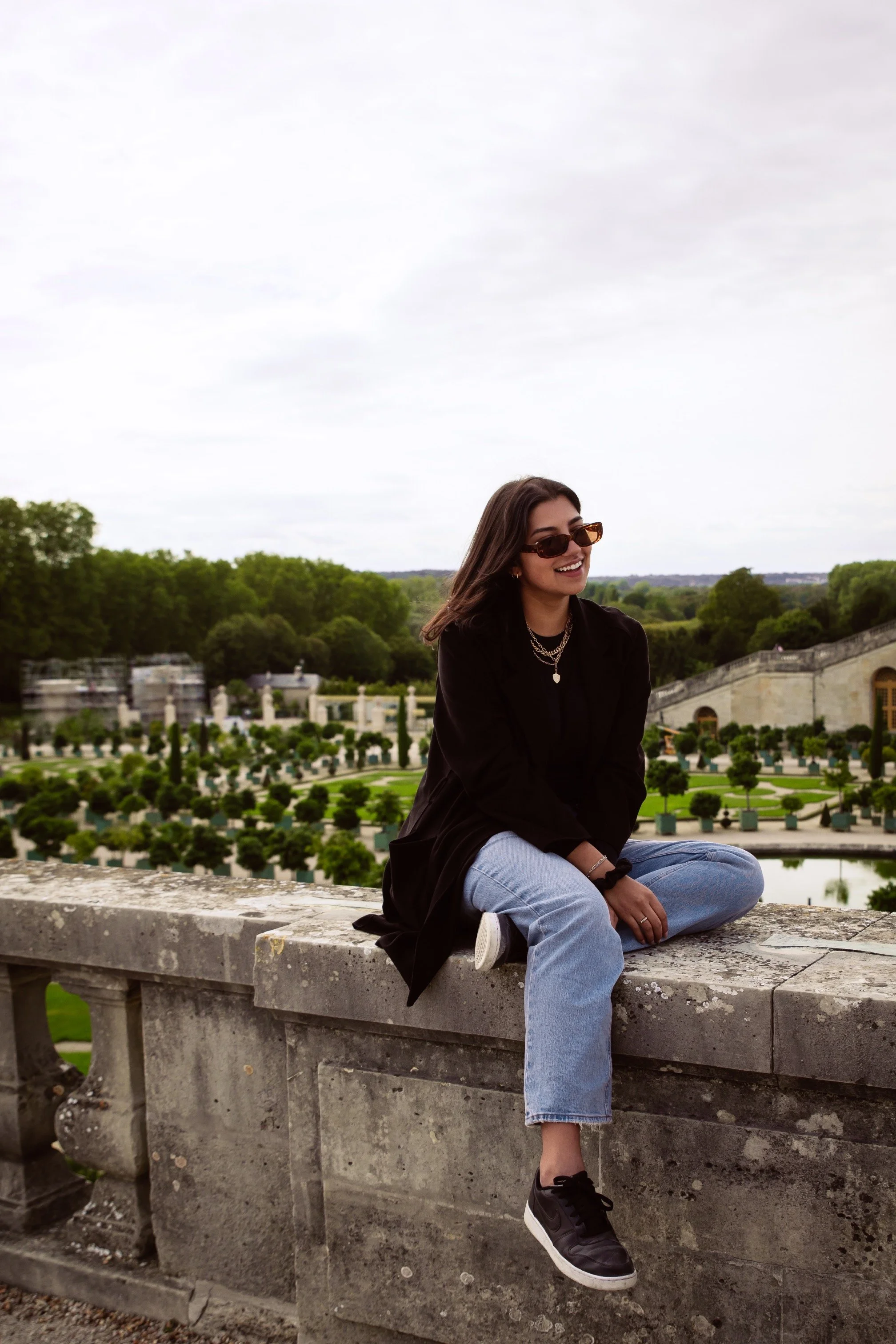 A young woman with dark hair, sunglasses, black jacket, light blue jeans, and black sneakers sitting on a stone wall outdoors, smiling with a decorative garden with trimmed trees and classical architecture in the background.