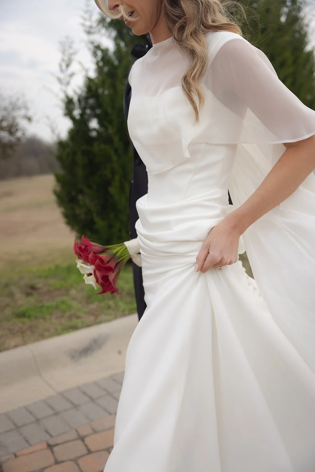 Close-up of a bride in a white wedding dress holding a small bouquet of red and white flowers, standing outdoors.