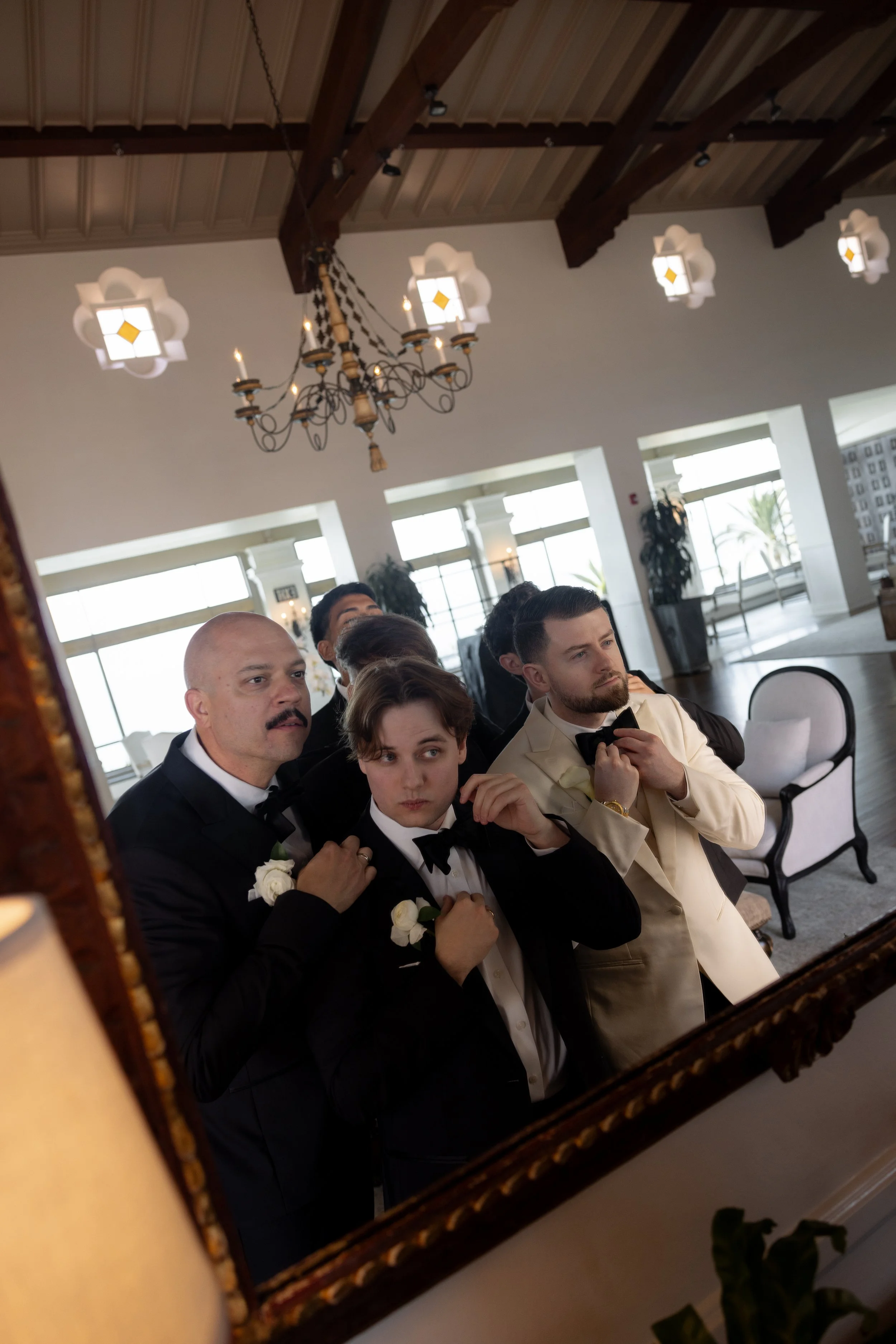 Group of men dressed in tuxedos looking into a mirror, adjusting their bow ties, fun groomsman pose