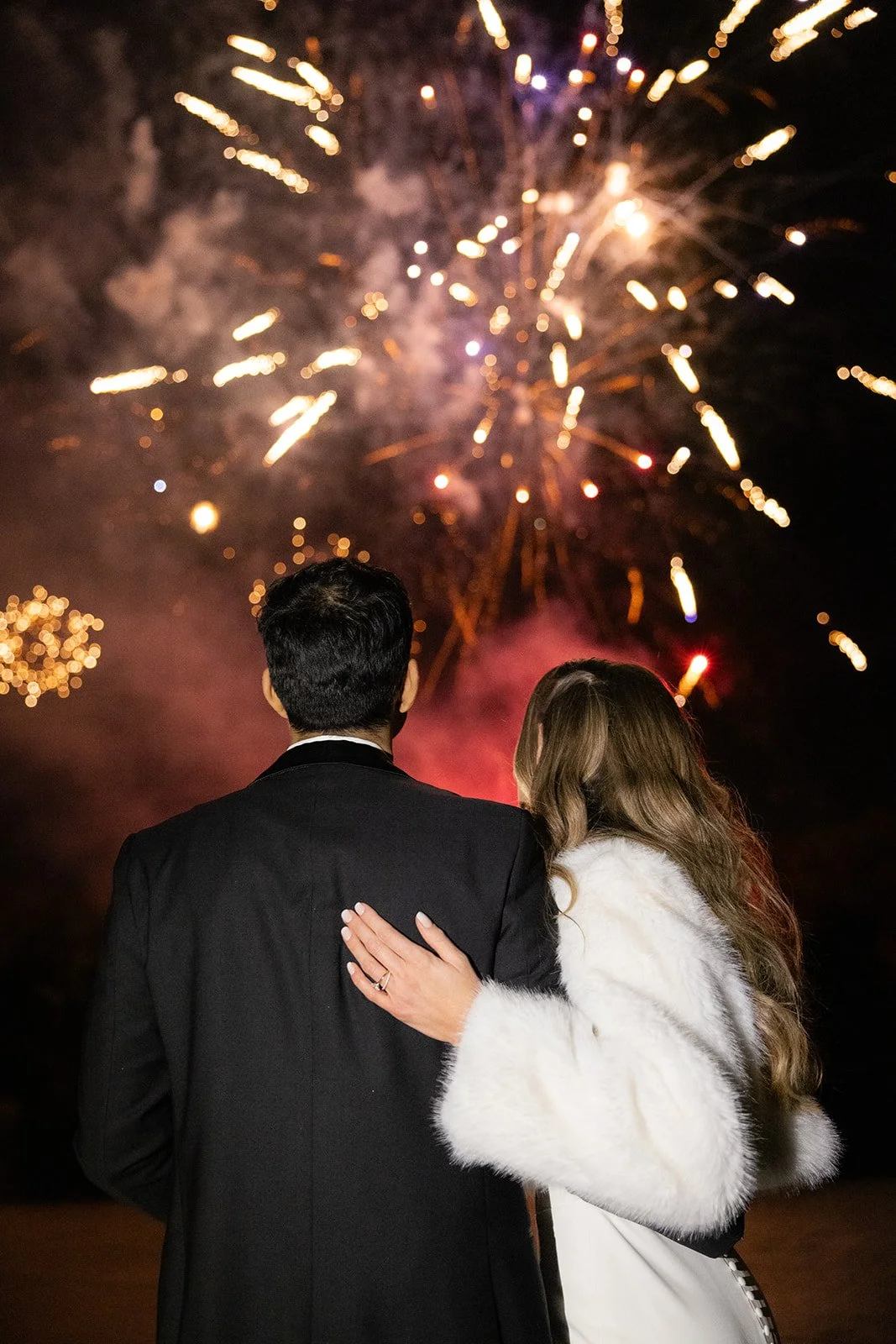 Couple watching fireworks at night, woman with her arm around man's shoulder, woman wearing a white fur coat, man in a suit.