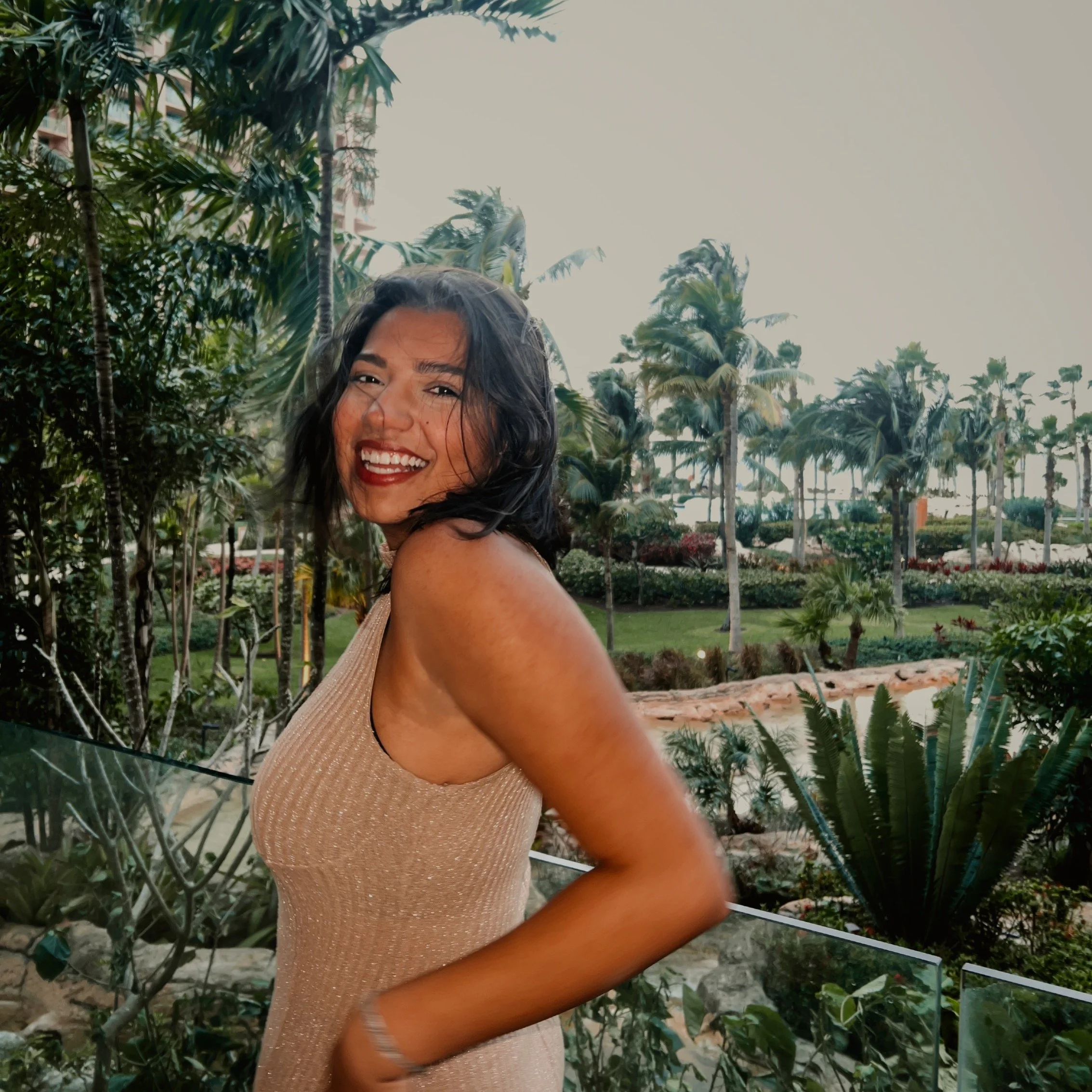 A woman smiling and posing outdoors in a tropical garden with palm trees and lush greenery.