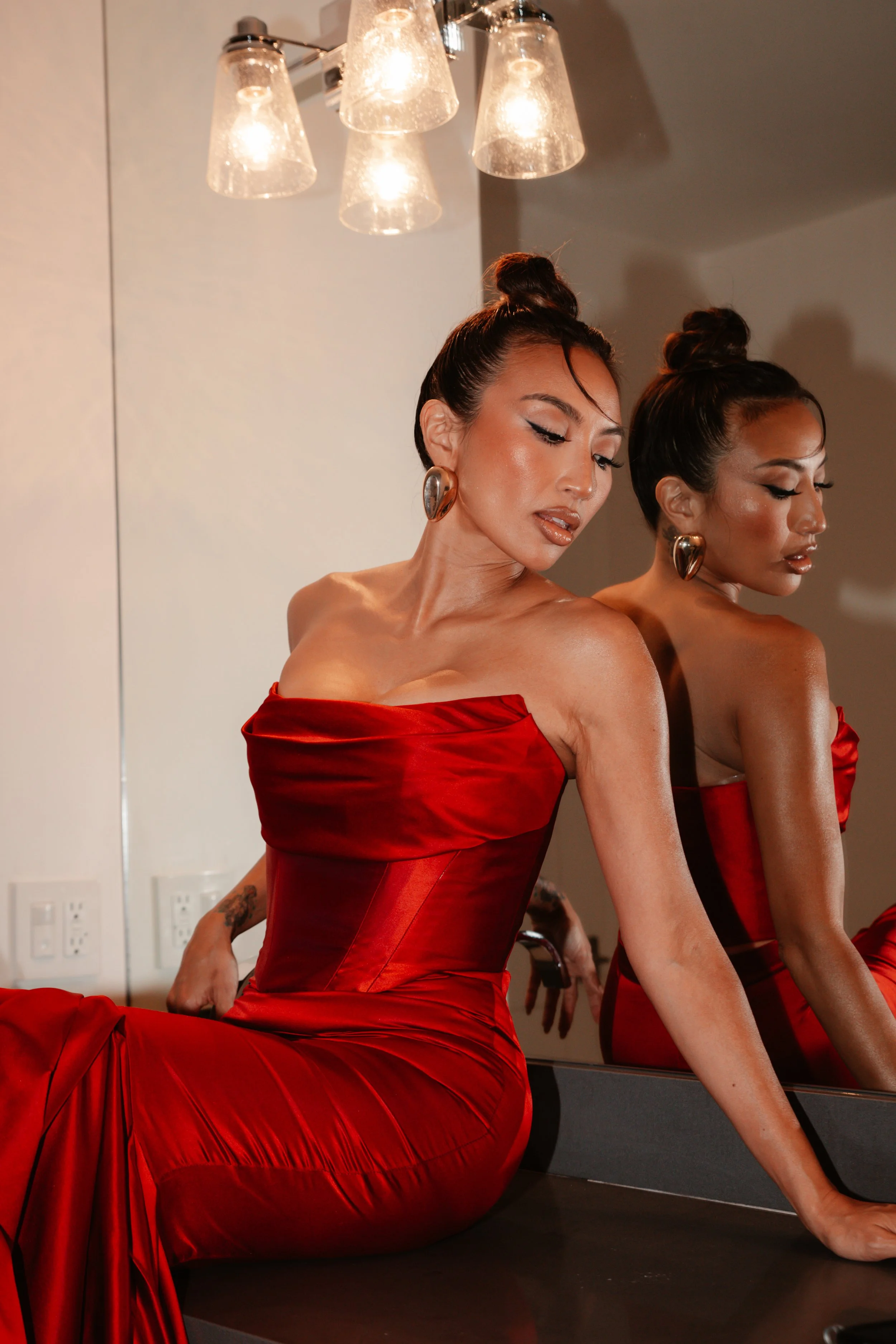 A woman in a red gown sitting in front of a mirror with her reflection visible. She has dark hair styled in an updo and is wearing large earrings. The room has a light fixture with multiple bulbs hanging from the ceiling.