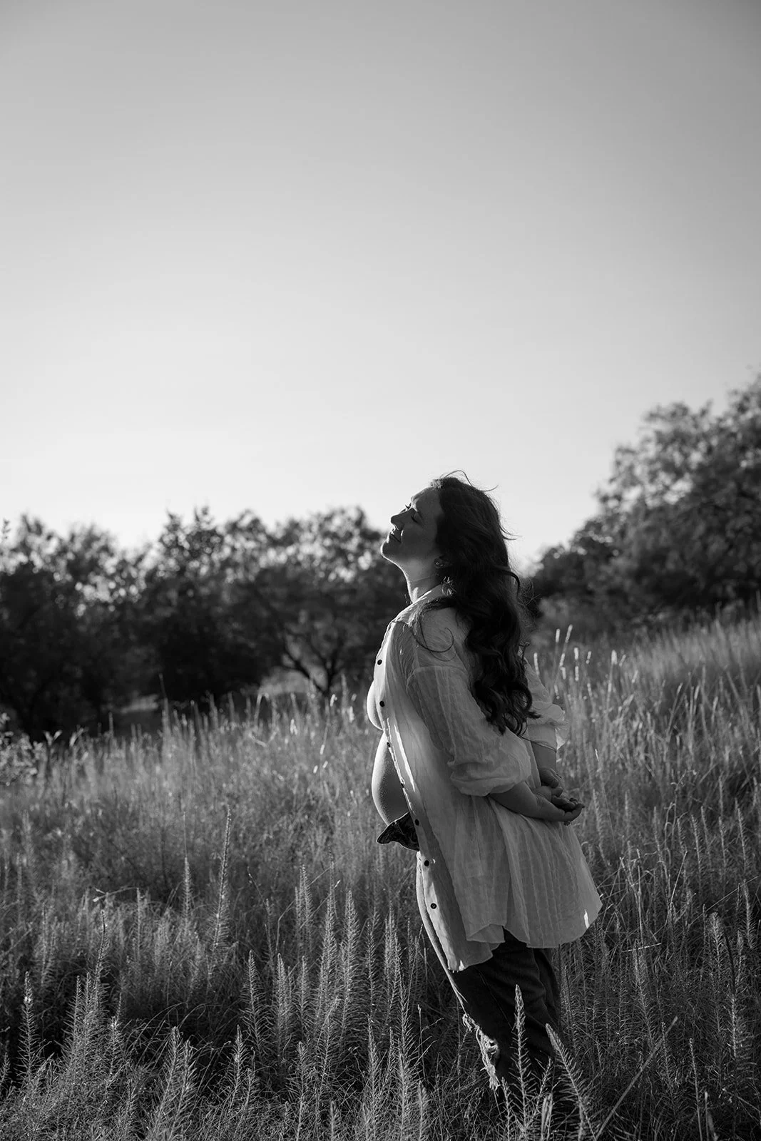 A pregnant woman standing in a field of tall grass with trees in the background, facing upwards with eyes closed in black and white.