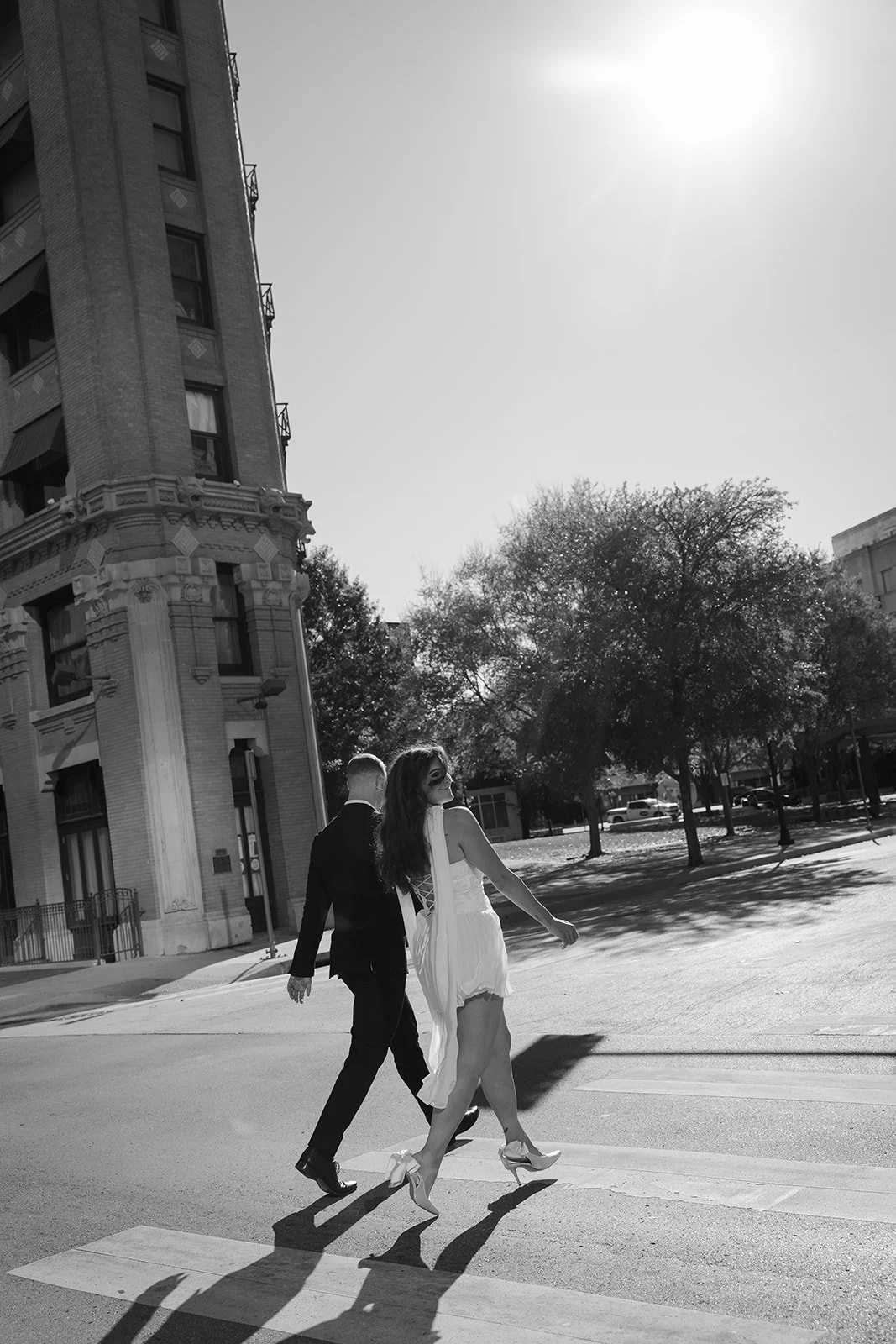 A black and white photo of a woman wearing a white dress and high heels, walking across a crosswalk with a man in a suit, on a city street with. Elopement Photo in Fort Worth Texas