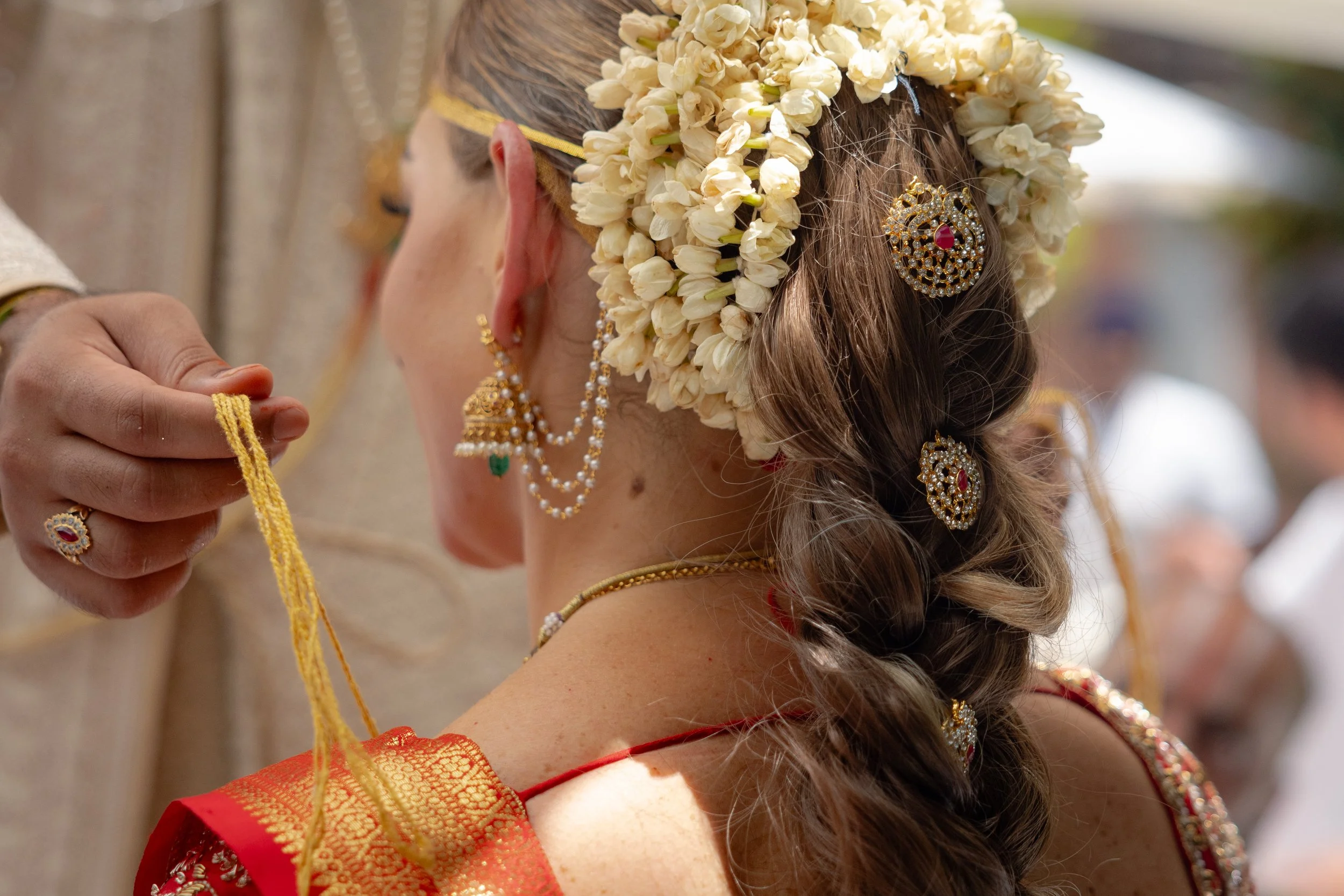 A woman dressed in traditional Indian attire with floral jewelry, being adorned with a gold necklace by another person. Her hair is decorated with jasmine flowers and gold jewelry.
