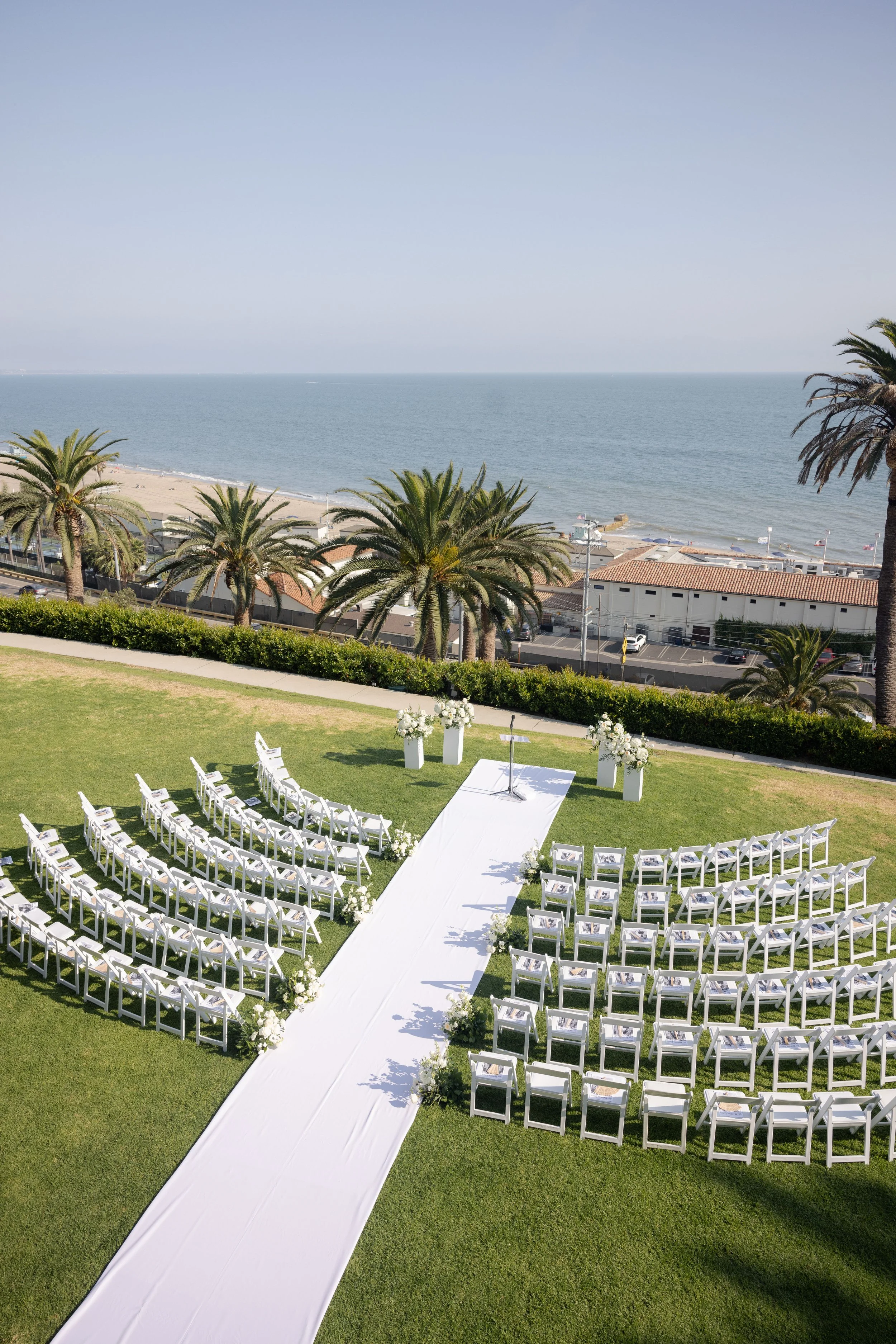 Outdoor wedding setup with white chairs on a grassy lawn, an aisle with white cloth, and a view of the ocean with palm trees in the background.