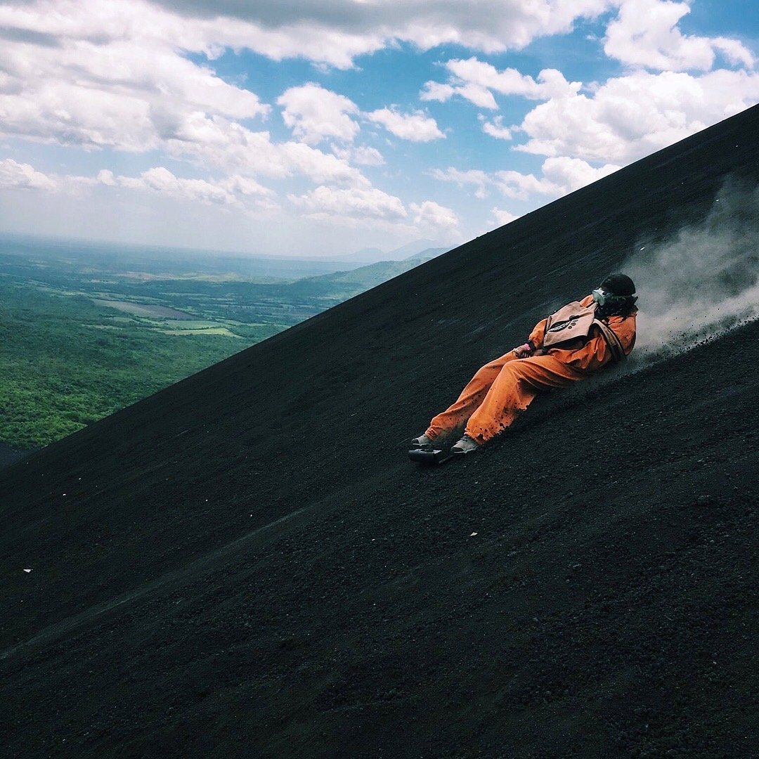 A person in an orange suit and helmet sliding down a dark volcanic slope, kicking up dust with a scenic landscape of green fields and a cloudy sky in the background.