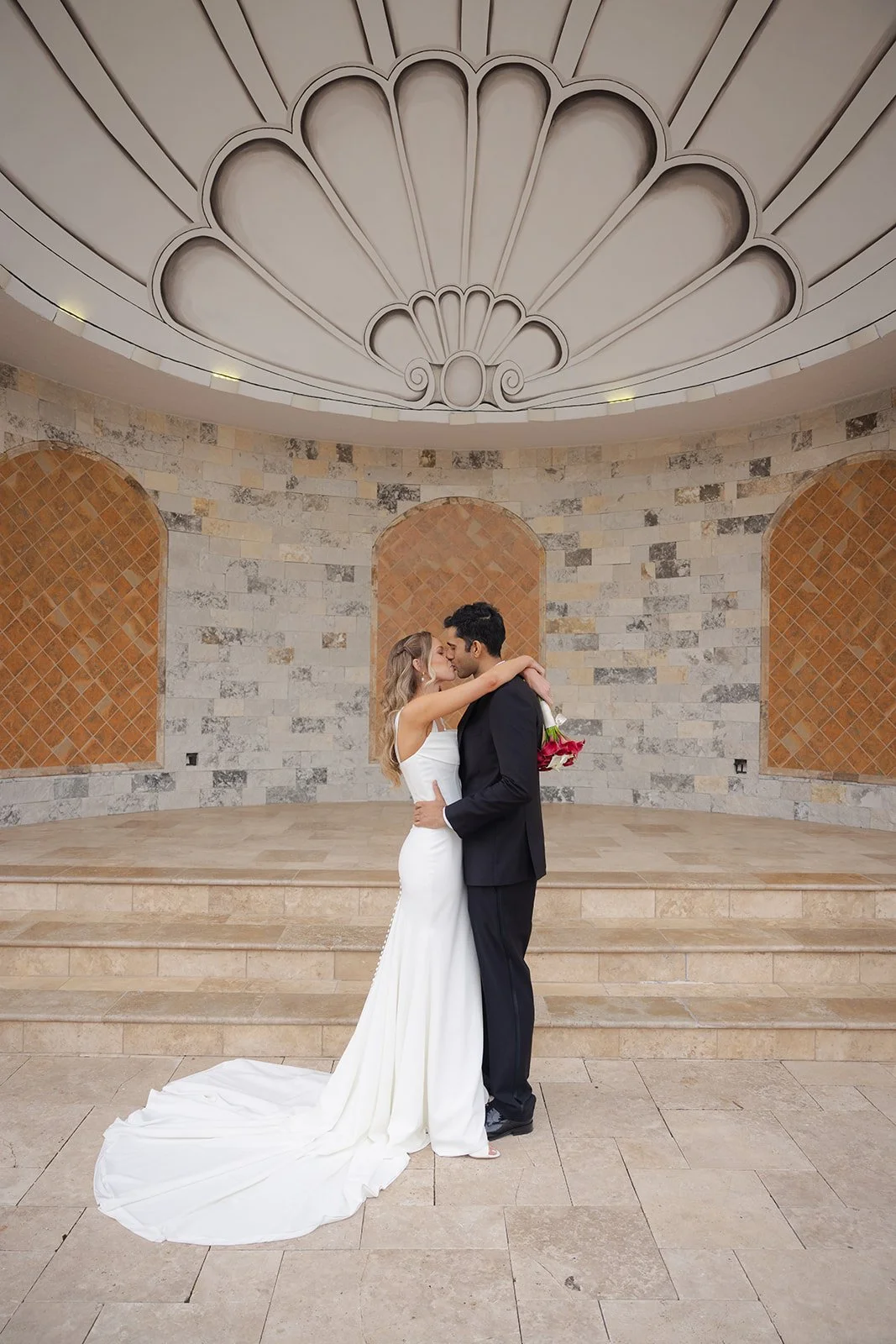 A bride and groom embrace and kiss in a wedding photo inside a decorated venue with arched brick and stone walls and a large decorative ceiling.