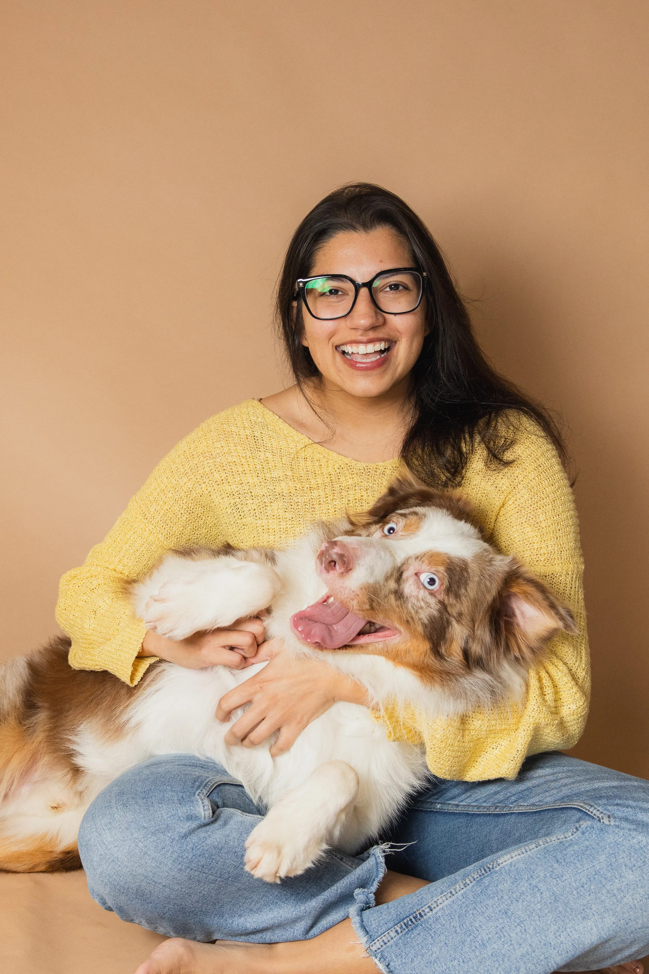 A smiling woman with glasses wearing a yellow sweater sitting on the floor, holding and playing with an Australian Shepherd dog with blue eyes against a beige background.