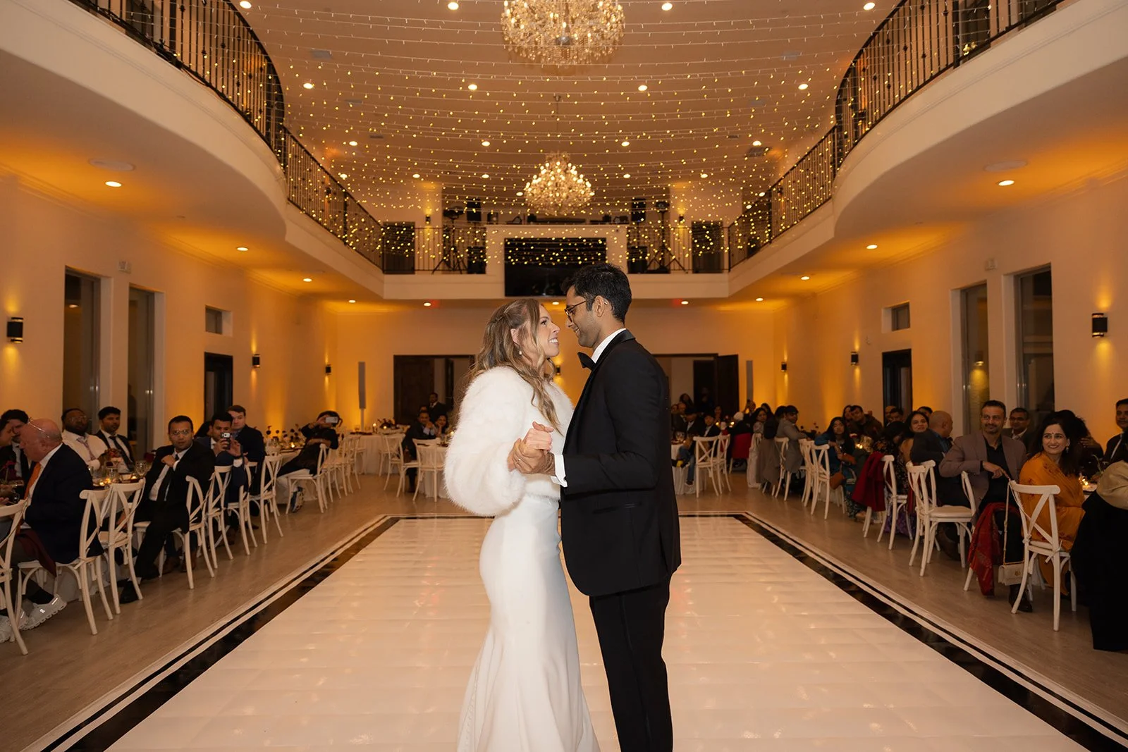 A bride and groom share their first dance in a wedding reception hall decorated with string lights and chandeliers, with seated guests watching.