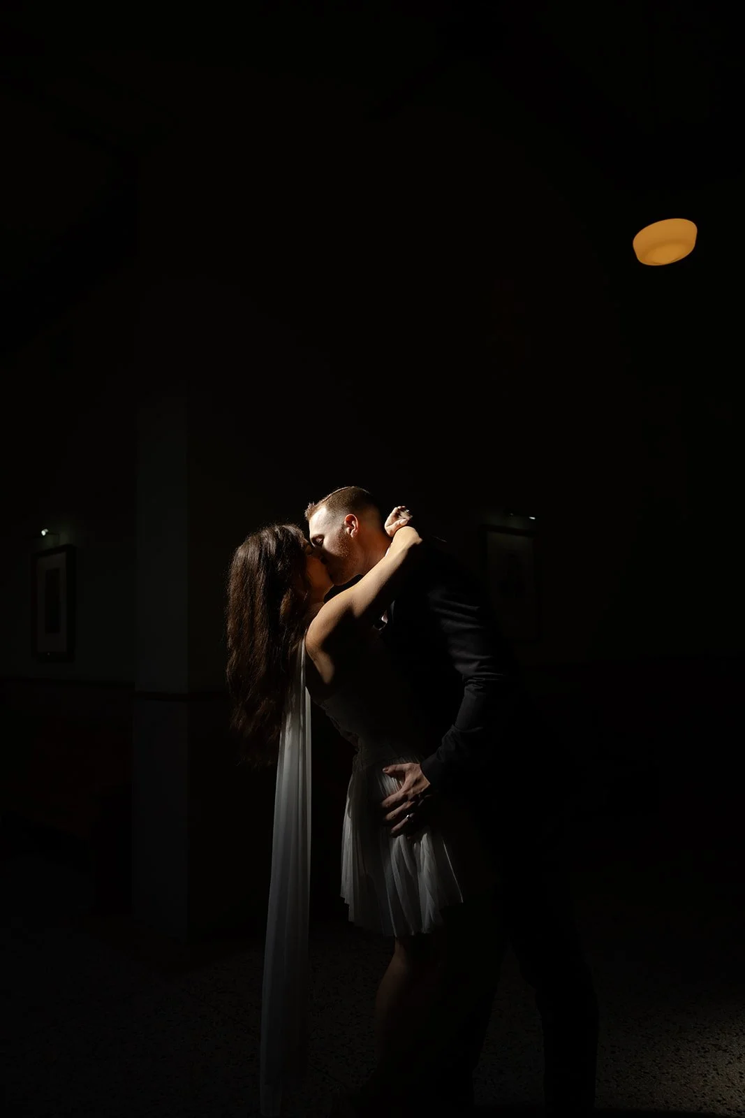 A couple kissing in a dimly lit room with minimal lighting and shadows.