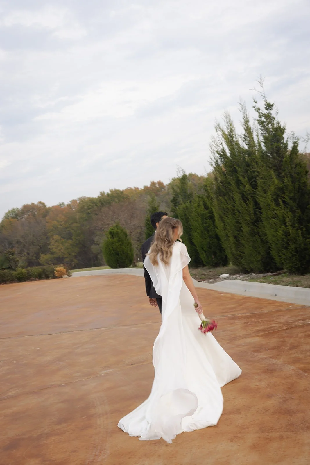 A bride in a white wedding dress holding a bouquet of pink flowers walking outdoors with a person in black. Green trees and an overcast sky in the background. Editorial style wedding image