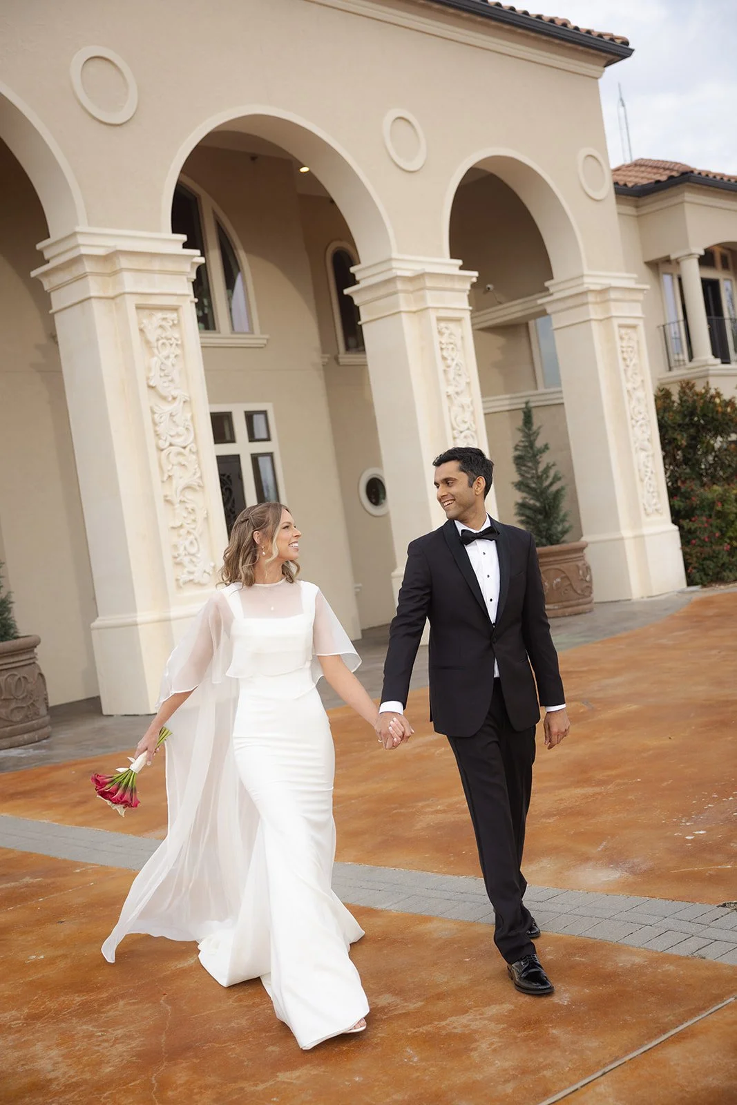 A newly married couple walking hand in hand outside a mansion. The bride is wearing a white dress and holding a bouquet of pink flowers, and the groom is in a black tuxedo. Walking shot of newlywed couple