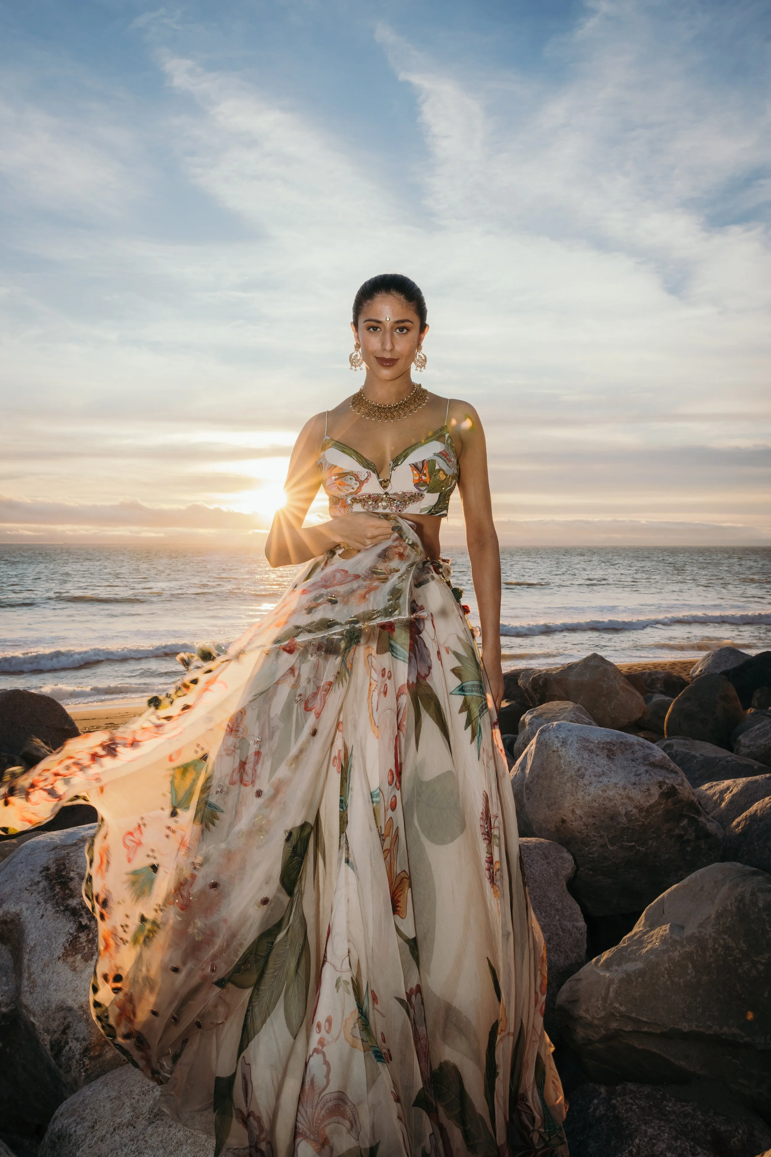 A woman wearing a floral dress standing on rocks at the beach during sunset.