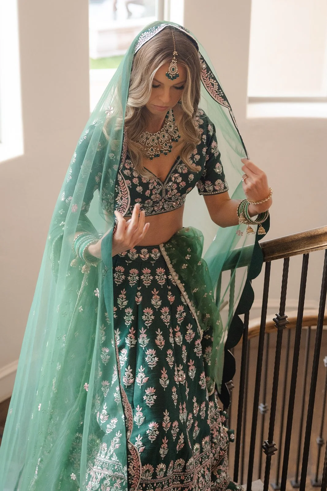 A woman dressed in traditional Indian attire, standing on a staircase indoors, wearing a dark embroidered lehenga with pink and silver flower patterns, a matching blouse, and a sheer green dupatta with embroidered edges, decorated with jewelry includ