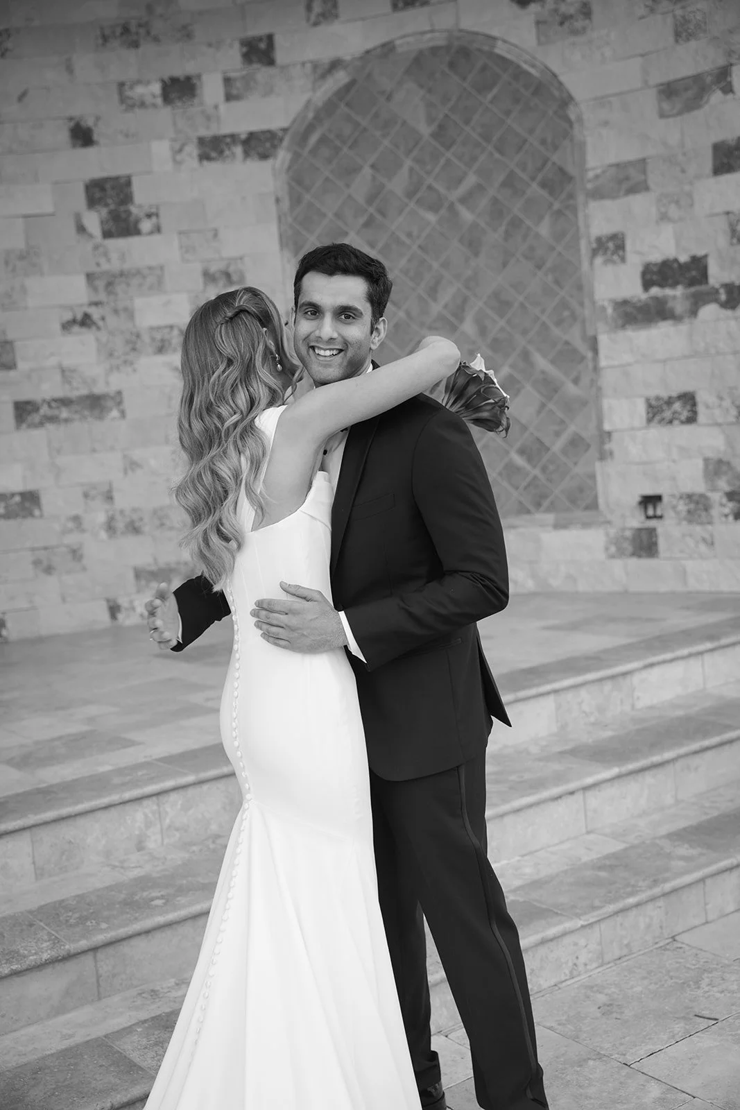 A smiling couple dancing, with the woman wearing a long white gown and the man in a dark suit, on a stone outdoor staircase with a brick wall backdrop.