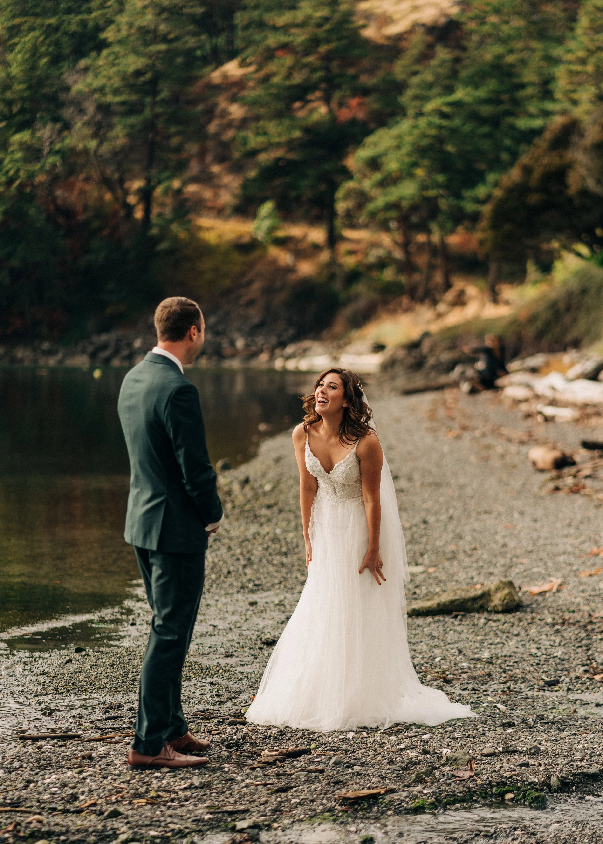A bride in a white wedding dress smiling and laughing while talking to a groom in a dark suit on a rocky shoreline surrounded by trees and a body of water.