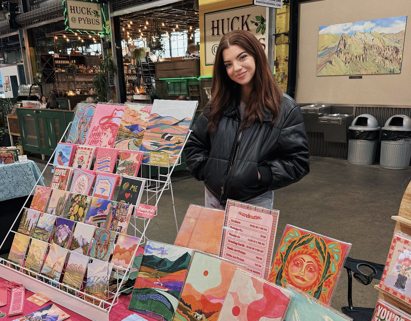 Young woman with long brown hair wearing a black leather jacket standing behind a display of colorful greeting cards and artwork at an indoor market or store.
