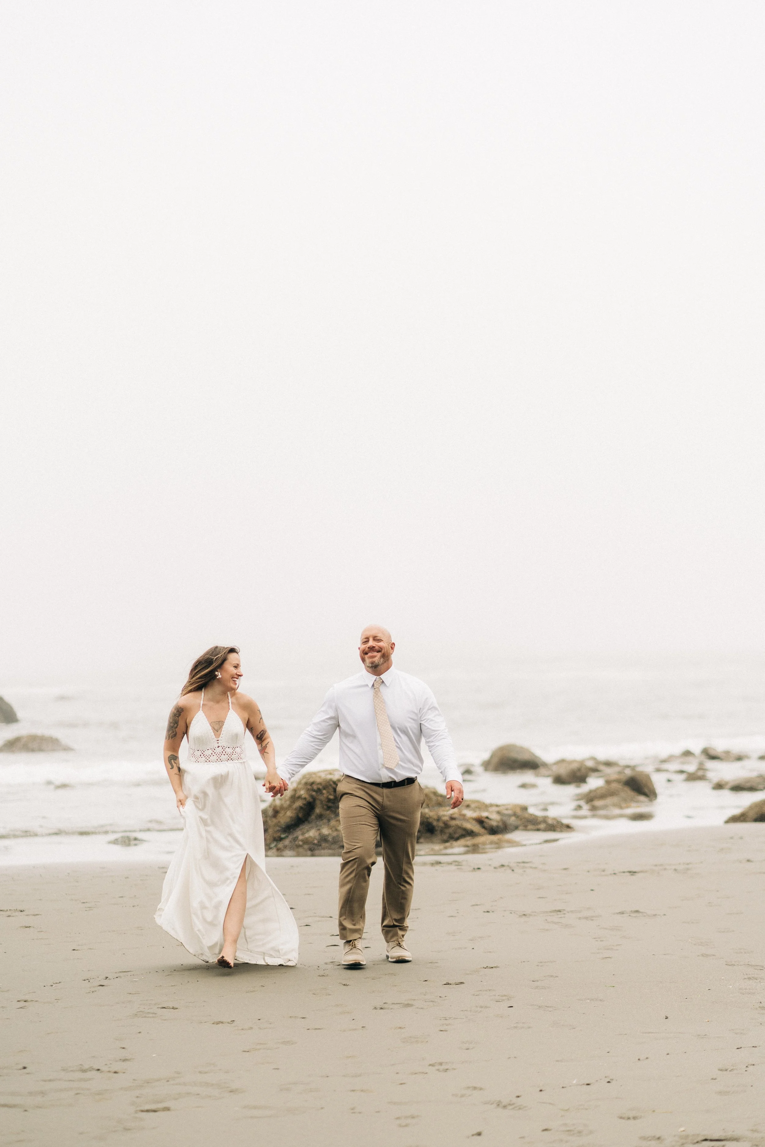 A couple dressed in wedding attire walking hand in hand on a beach, smiling and enjoying their time together.
