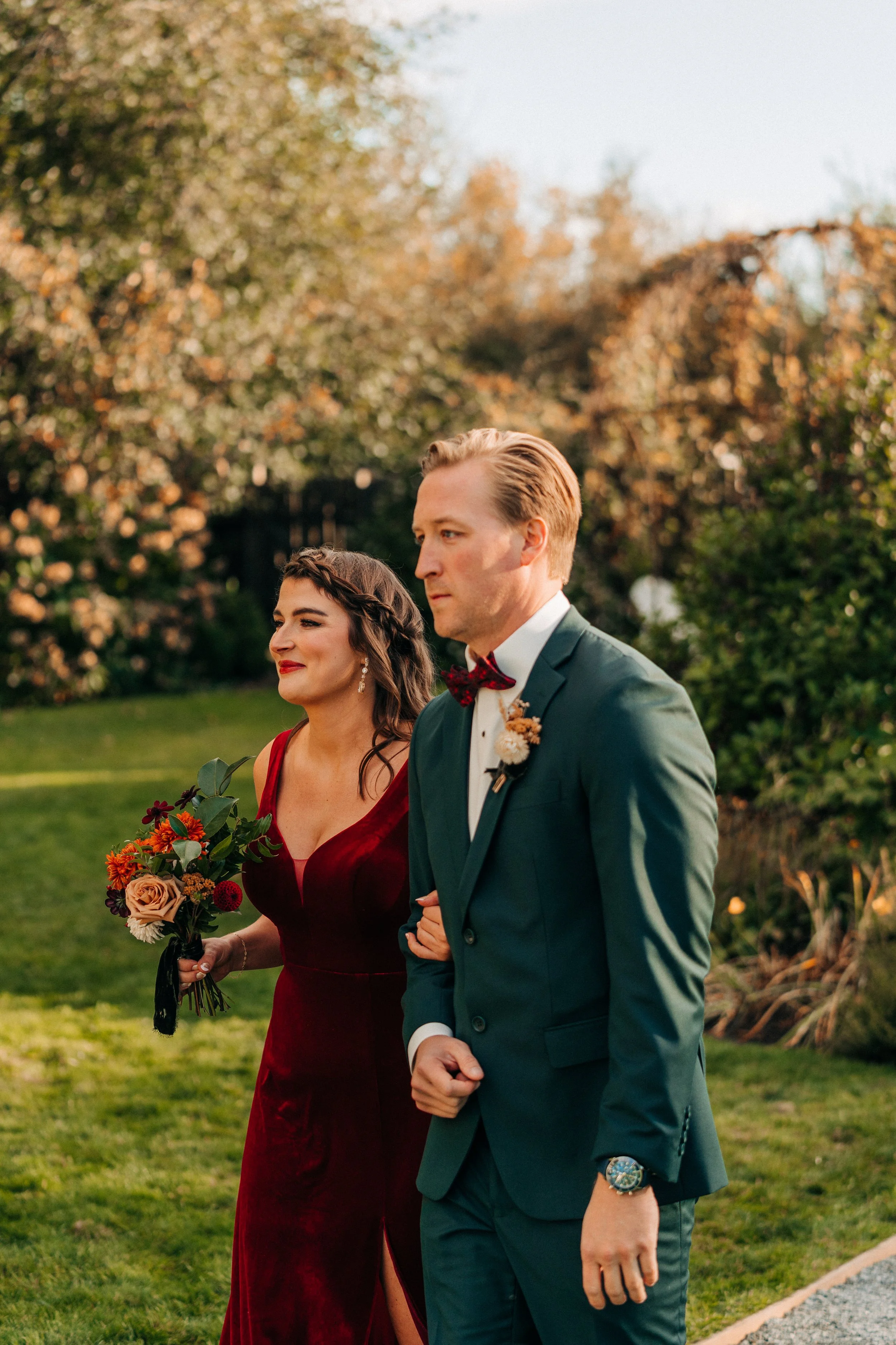 A man in a green suit and a woman in a red dress walking together outdoors, with the woman holding a bouquet of flowers.