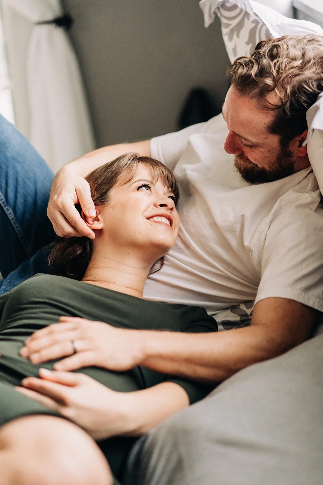 A woman and a man lying on a bed, smiling and looking into each other's eyes, in a cozy bedroom setting.